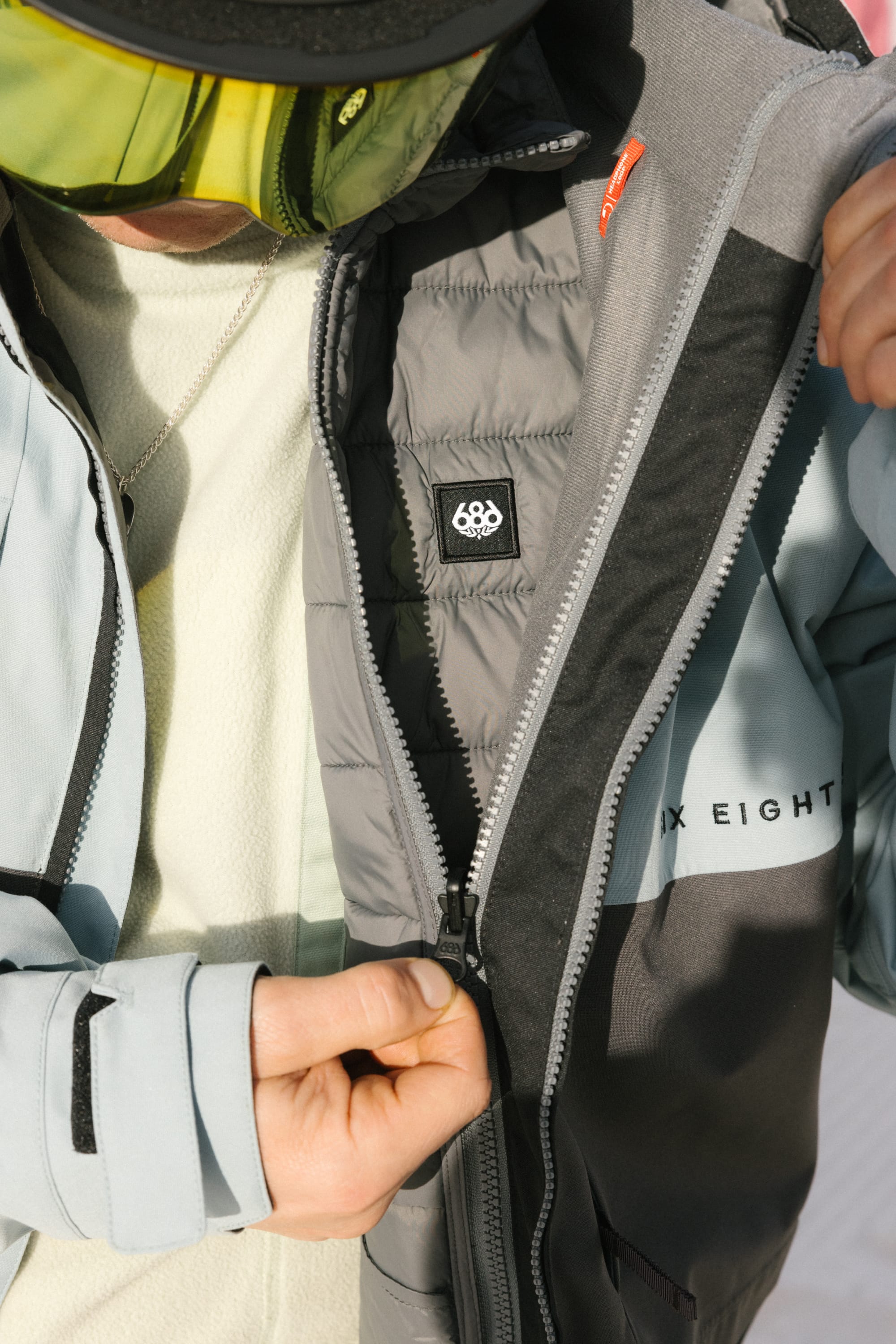 Wearing the 686 Mens Smarty 3-In-1 Form Jacket, a person zips up to reveal its quilted lining and signature 686 logo. Yellow ski goggles cover their eyes; only their hands and part of their face are visible.