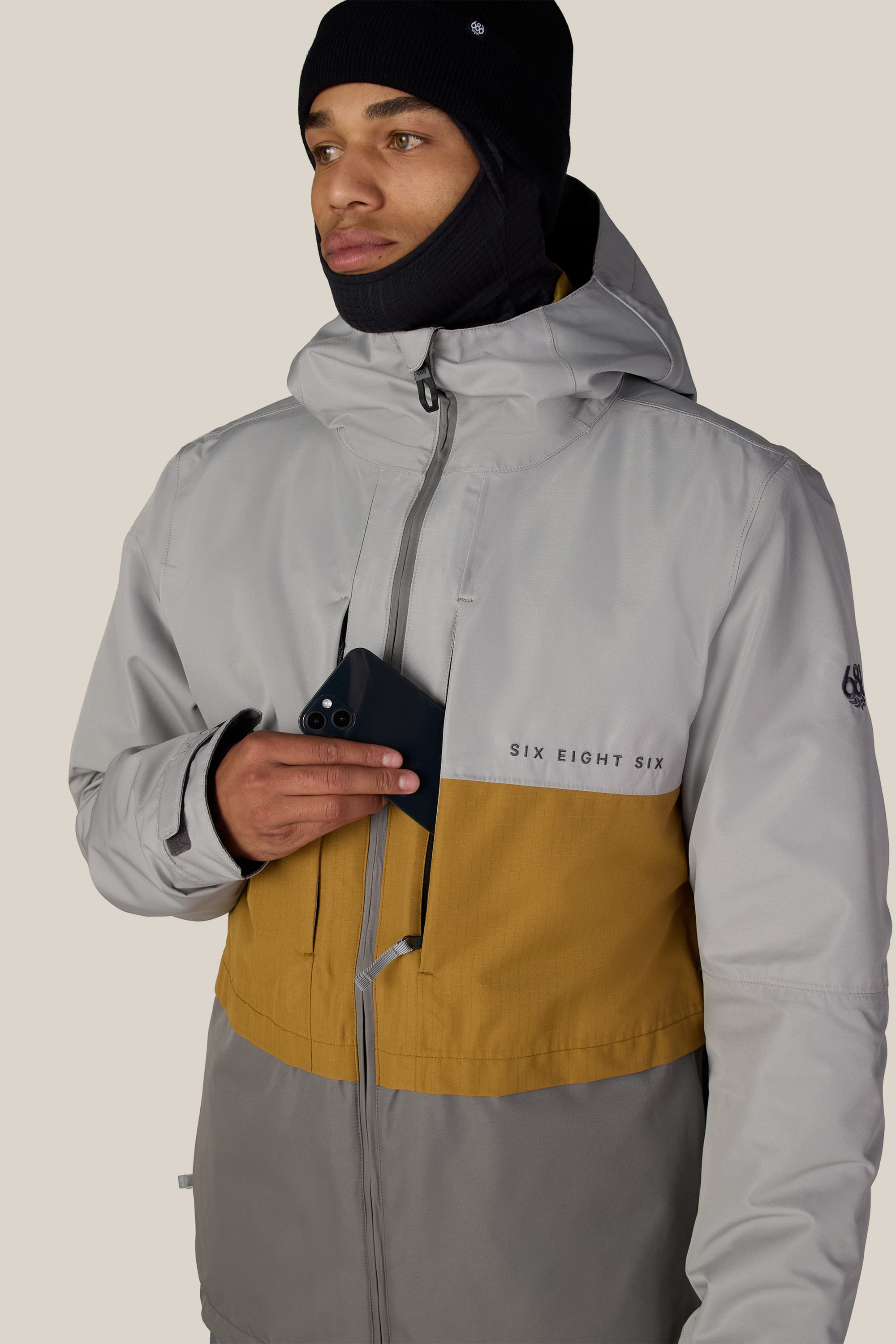 A person wearing the 686 Men’s SMARTY® 3-In-1 Form Jacket in gray and tan, along with a black beanie and face covering, holds a black smartphone partially inside the jacket’s chest pocket.