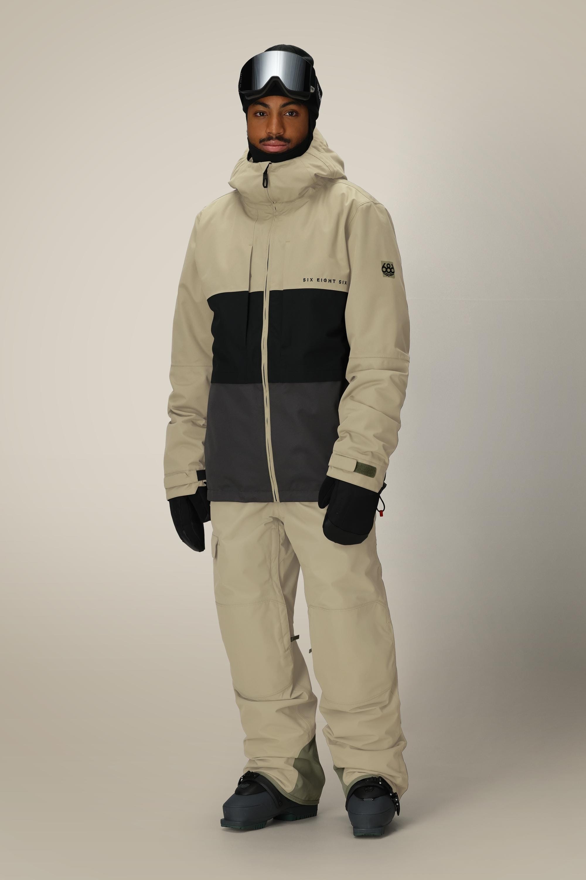 A person wearing the 686 Mens Smarty 3-In-1 Form Jacket in beige and black, paired with beige snow pants, black gloves, boots, and ski goggles stands against a plain light background.