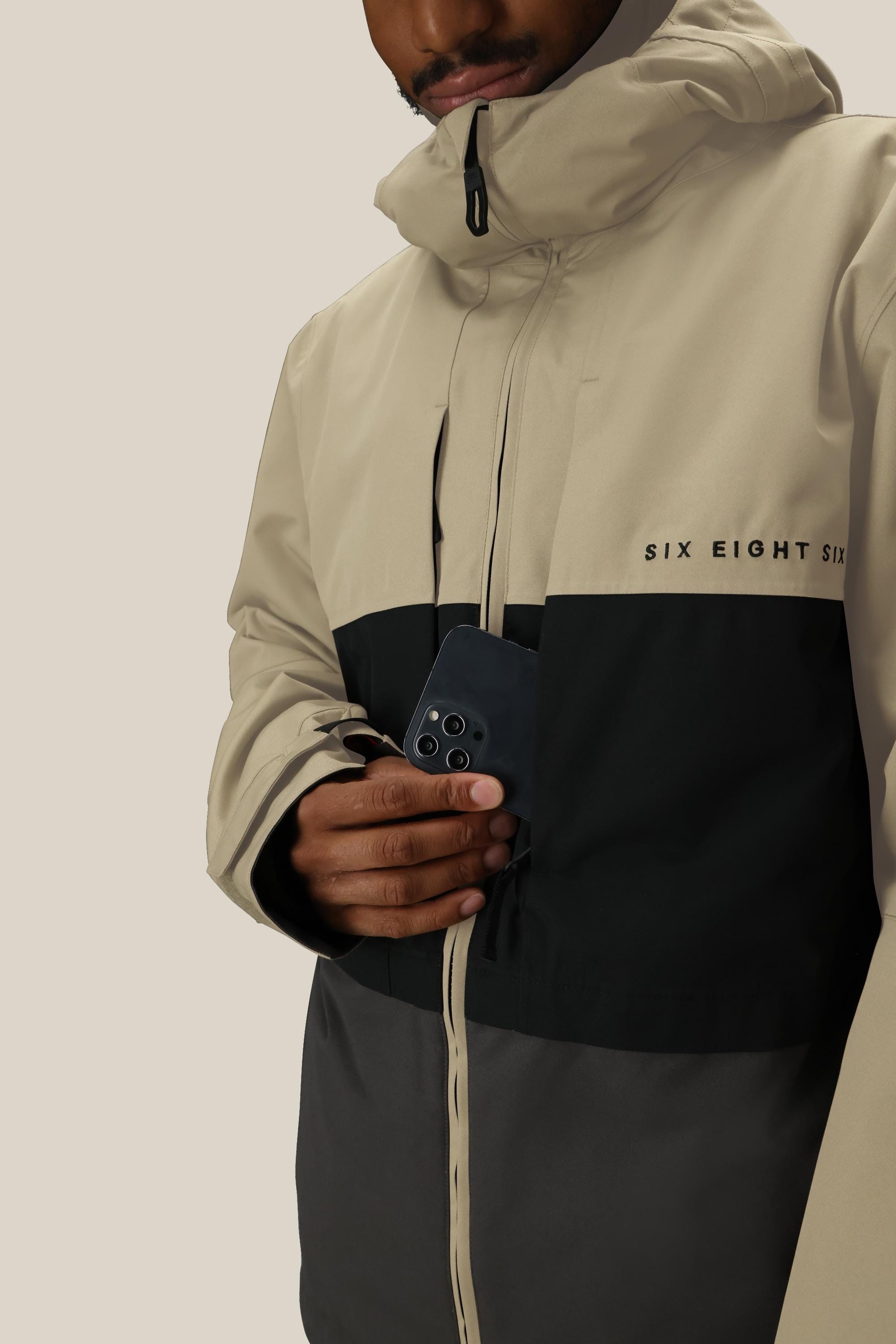 Wearing the 686 Mens Smarty 3-In-1 Form Jacket in beige and black, a person places a black smartphone into the zippered chest pocket, with the image cropped to show only their lower face and torso.