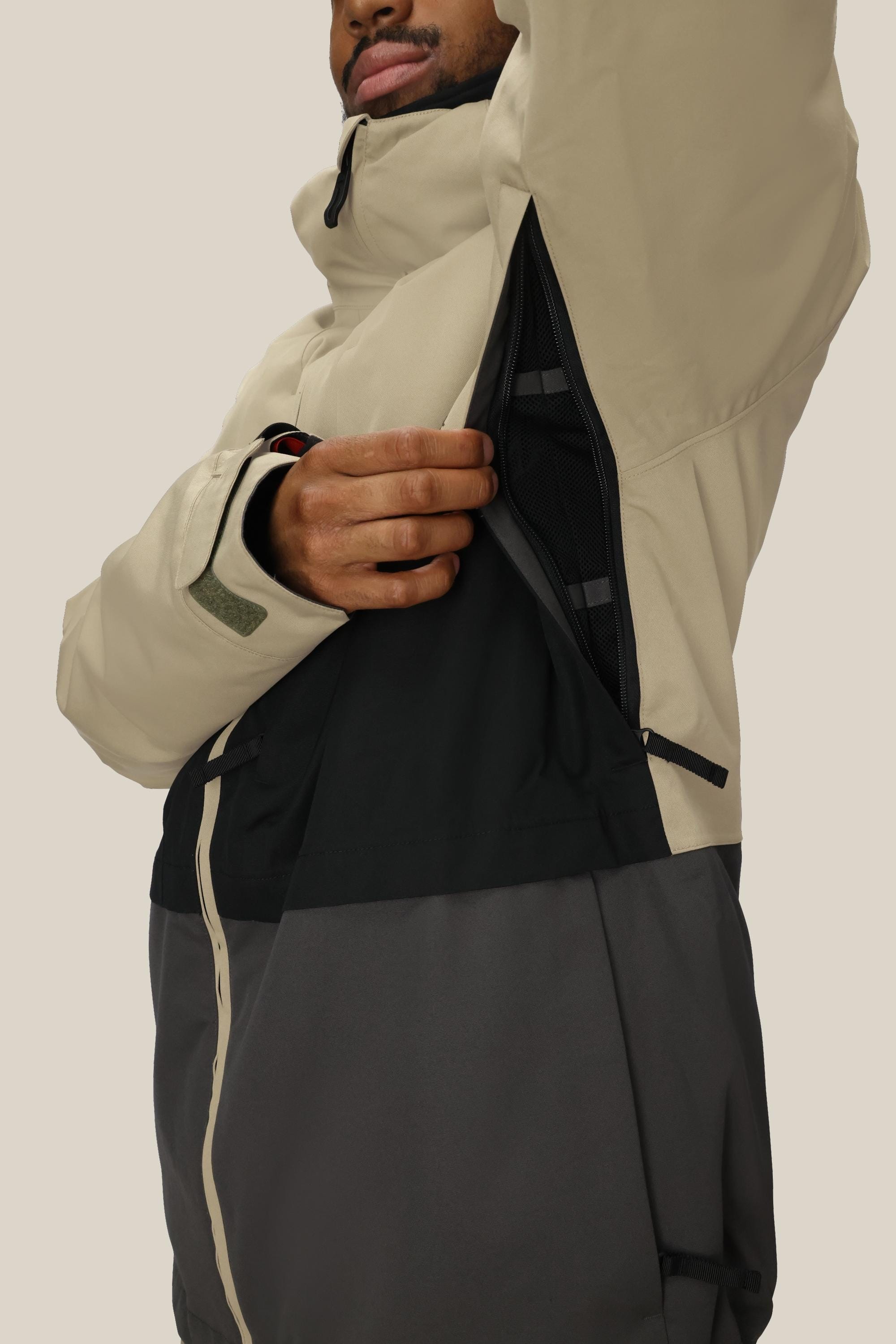 A man wearing the 686 Mens Smarty 3-In-1 Form Jacket in beige and black unzips a large underarm vent, revealing the mesh lining inside this jacket from 686.