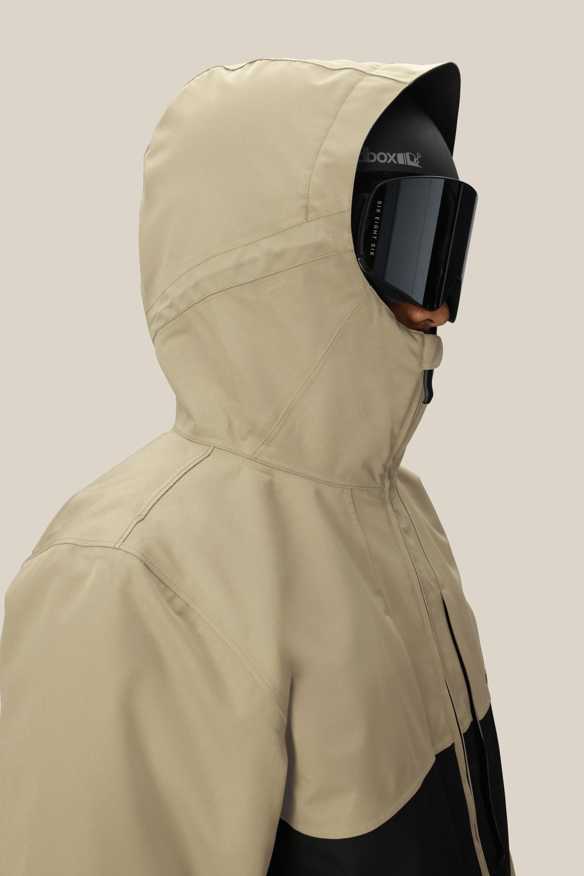 A person wearing the 686 Mens Smarty 3-In-1 Form Jacket in tan, paired with black ski goggles and a black face covering, is shown from the side against a plain beige background.