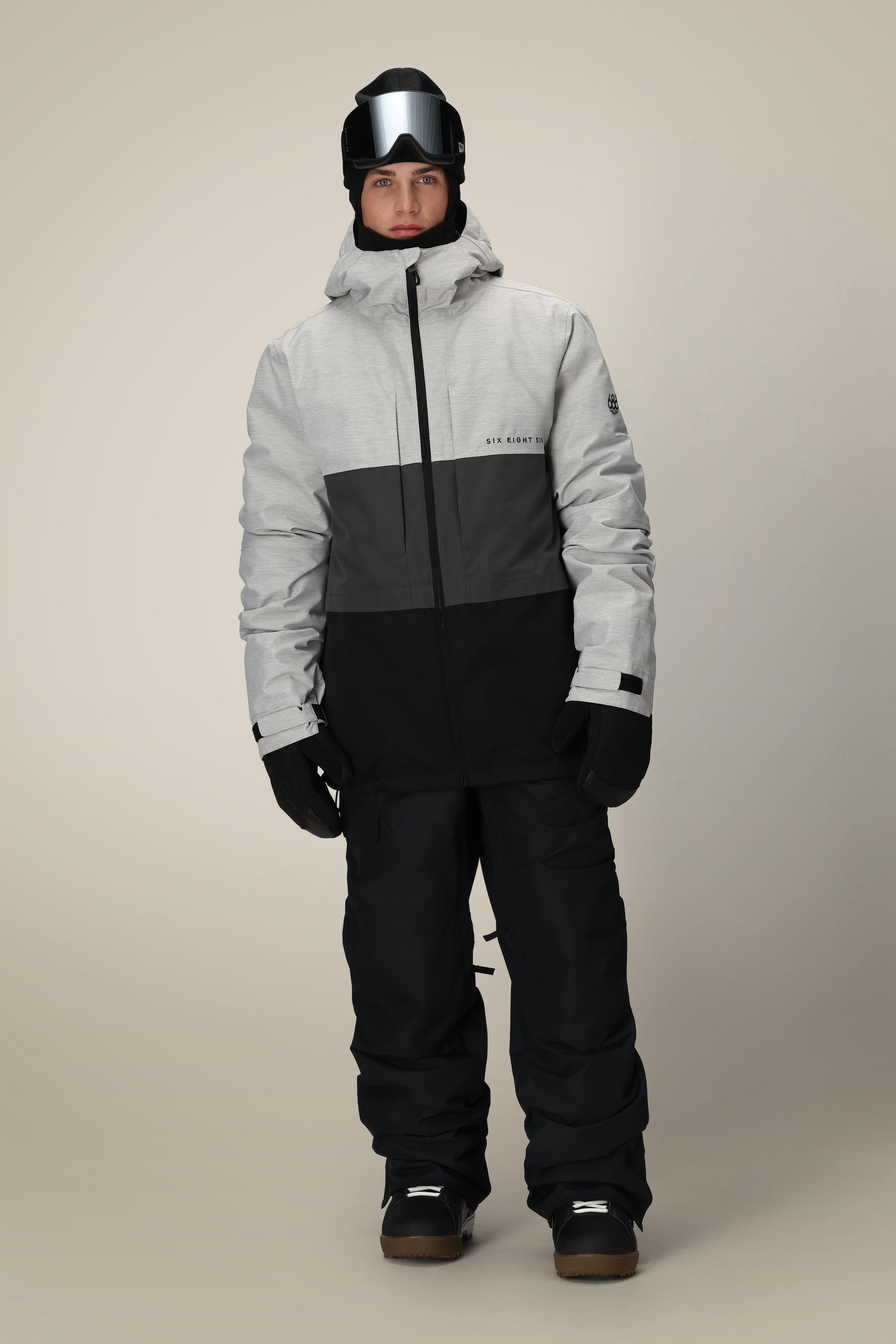 A man wearing a 686 Mens Smarty 3-In-1 Form Jacket in light gray and black, black snow pants, gloves, boots, a black helmet, and ski goggles stands facing forward against a plain beige background.