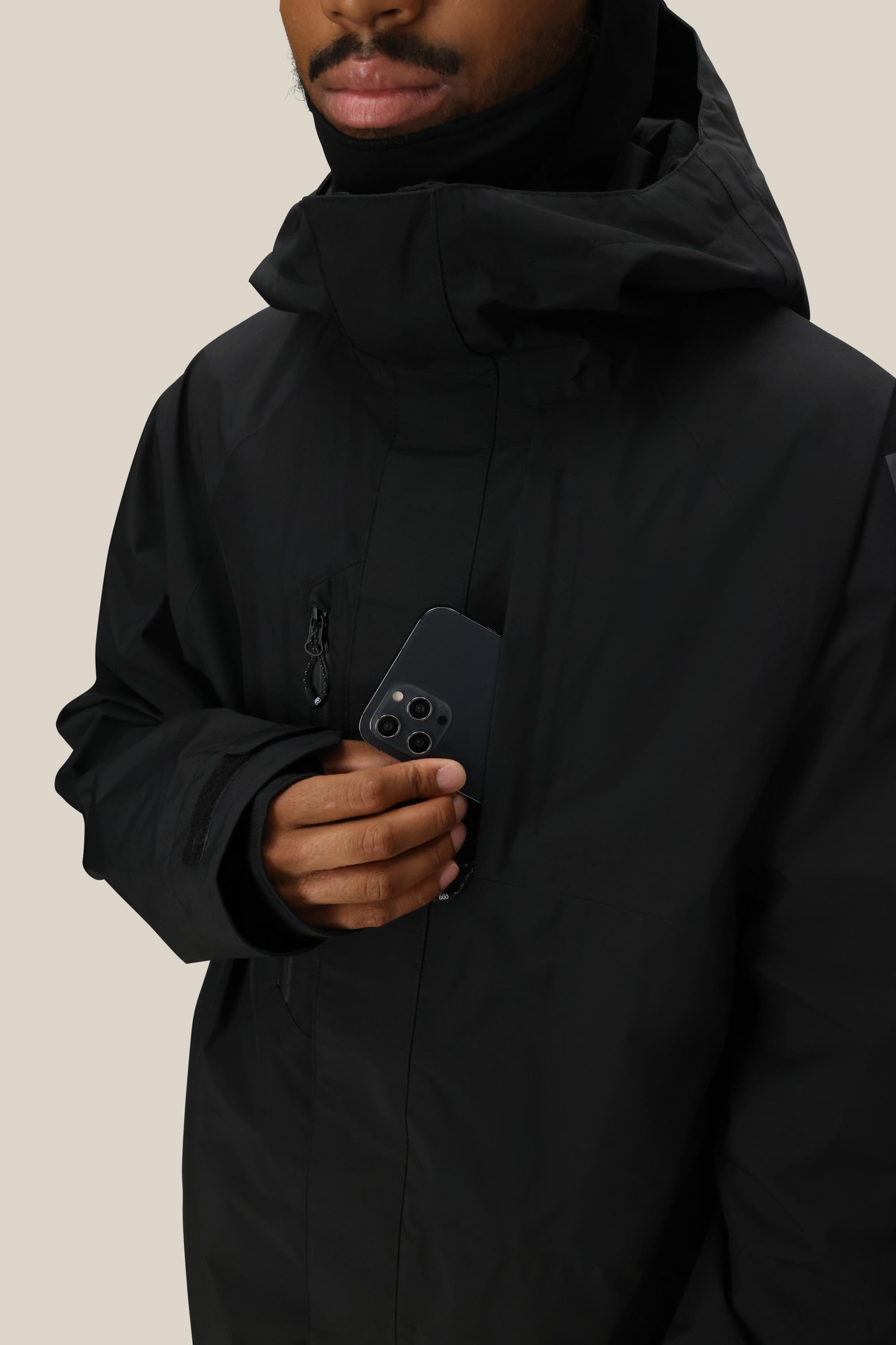 A person wearing the 686 Men’s GORE-TEX® Core Shell Jacket by 686 places a black smartphone with three cameras into the jacket’s zippered chest pocket, against a plain light background.