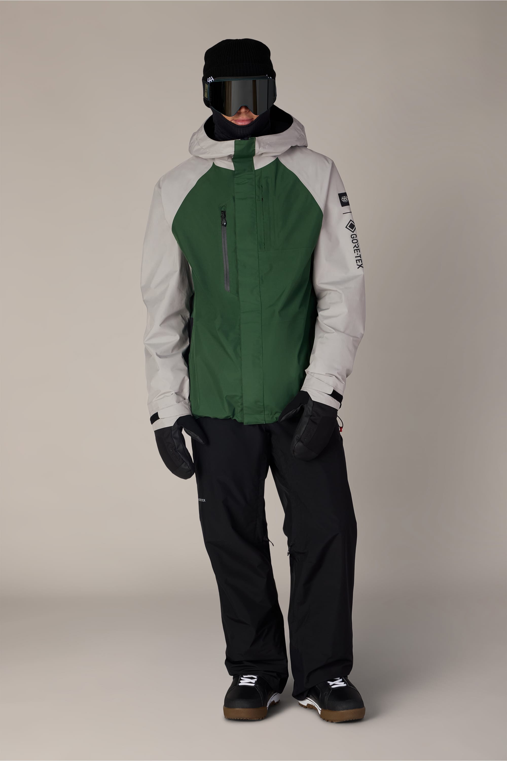 A person wearing the 686 Men’s GORE-TEX® Core Shell Jacket in green and gray, paired with black snow pants, black gloves, a face covering, large ski goggles, and black winter boots stands against a plain light background.