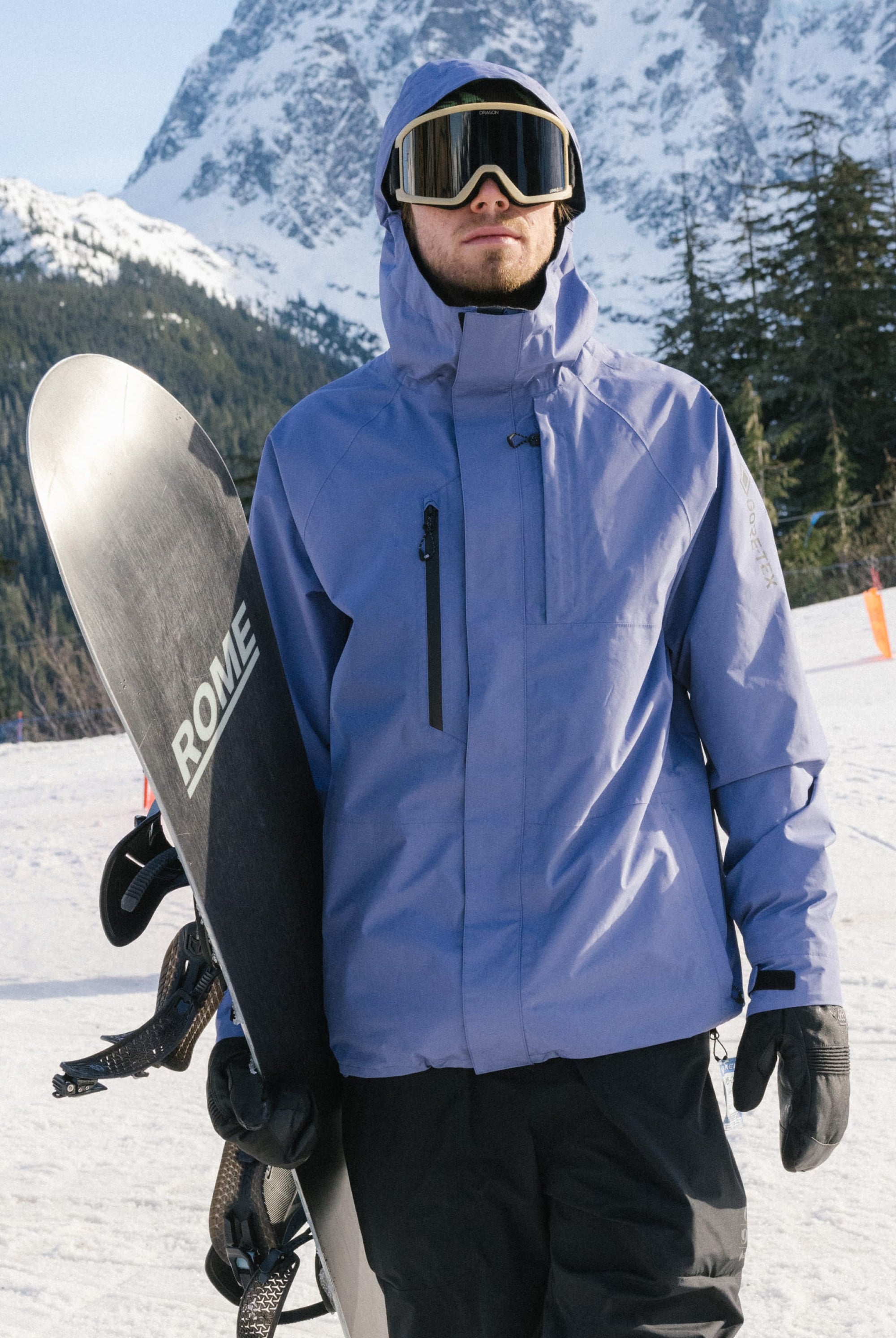 Wearing the 686 Mens GORE-TEX Core Shell Jacket in light purple, black pants, and ski goggles, a man stands on a snowy mountain holding a snowboard vertically with snow-covered trees and a mountain peak behind him.