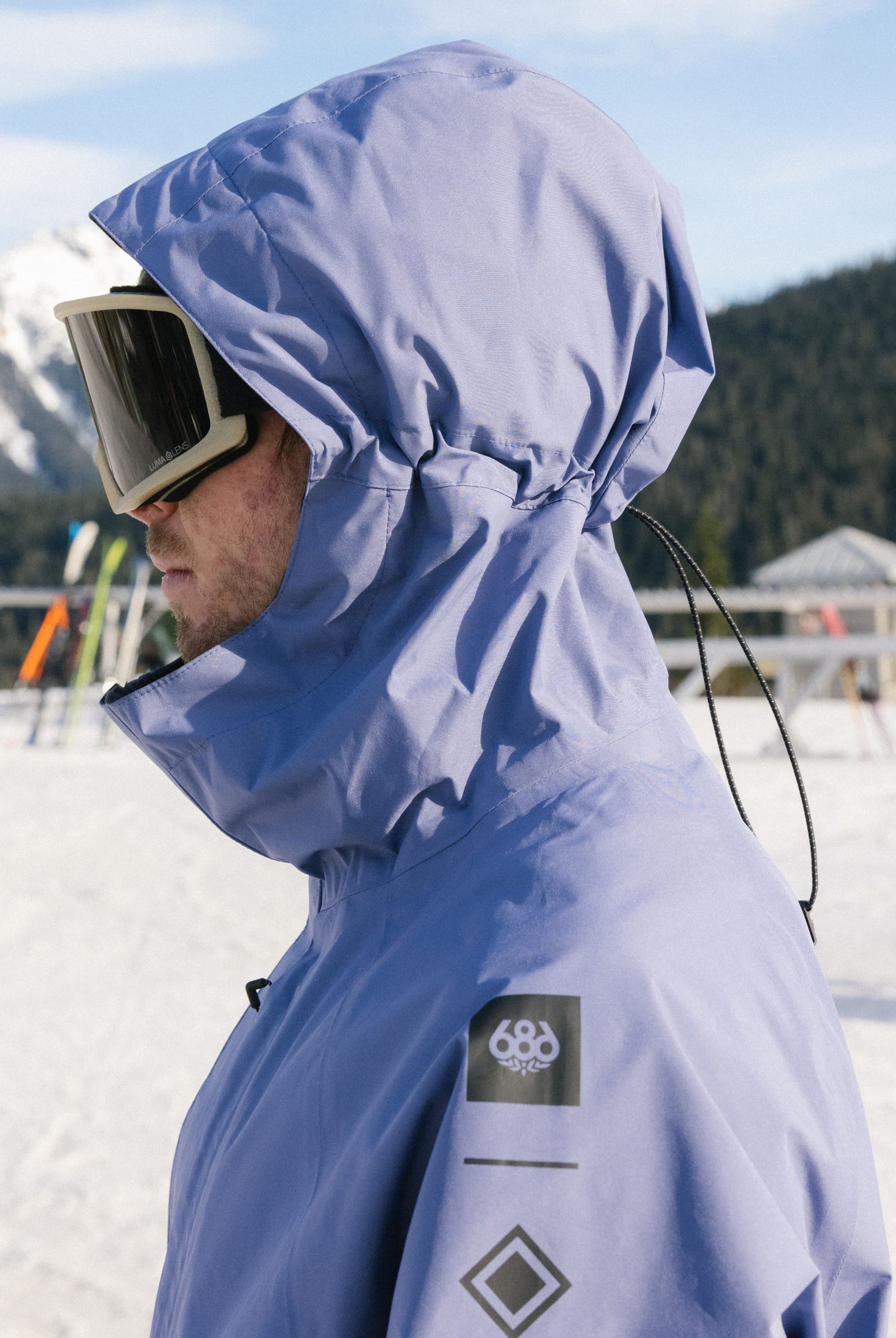 Wearing a 686 Mens GORE-TEX Core Shell Jacket and large ski goggles, a person stands outdoors on a snowy mountain with ski equipment and trees in the background.