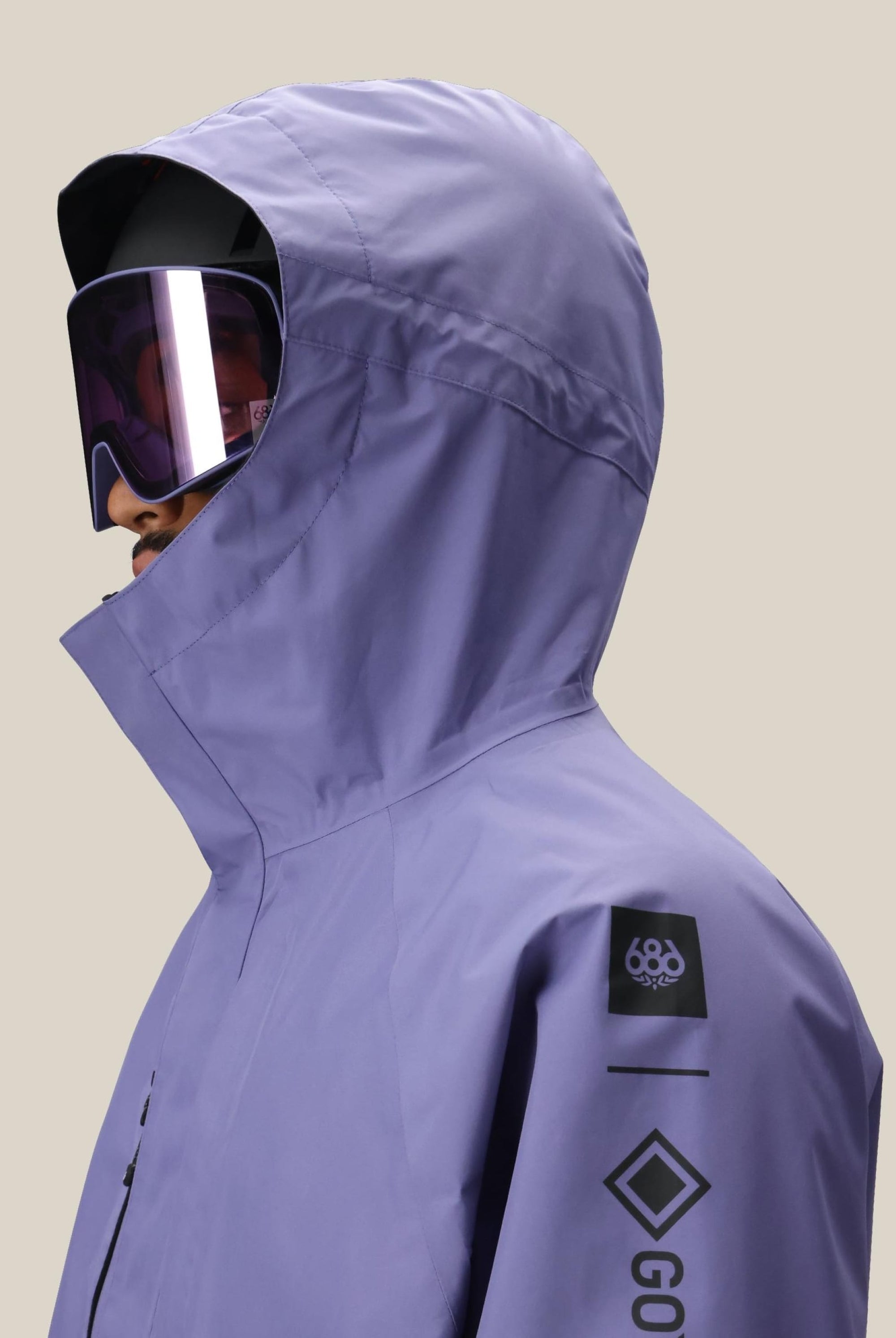 A person wearing the 686 Mens GORE-TEX Core Shell Jacket in light purple with black sleeve logos and large reflective ski goggles, facing left against a plain beige background.