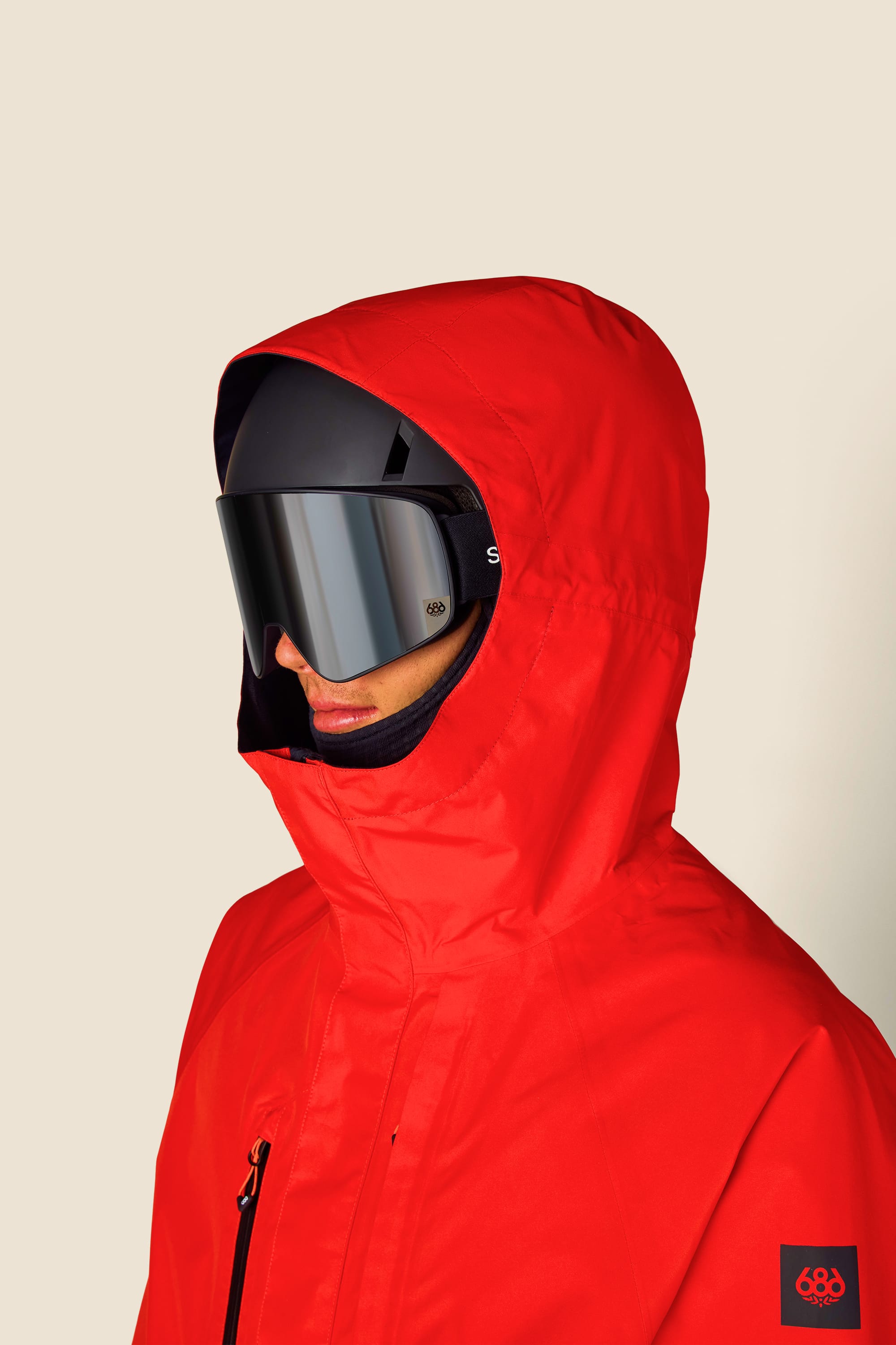 A person wearing the 686 Men’s GORE-TEX® Core Shell Jacket in bright red, paired with black ski goggles and a black helmet, is facing left. The background is plain light beige, with a small 686 logo featured in the lower right corner.