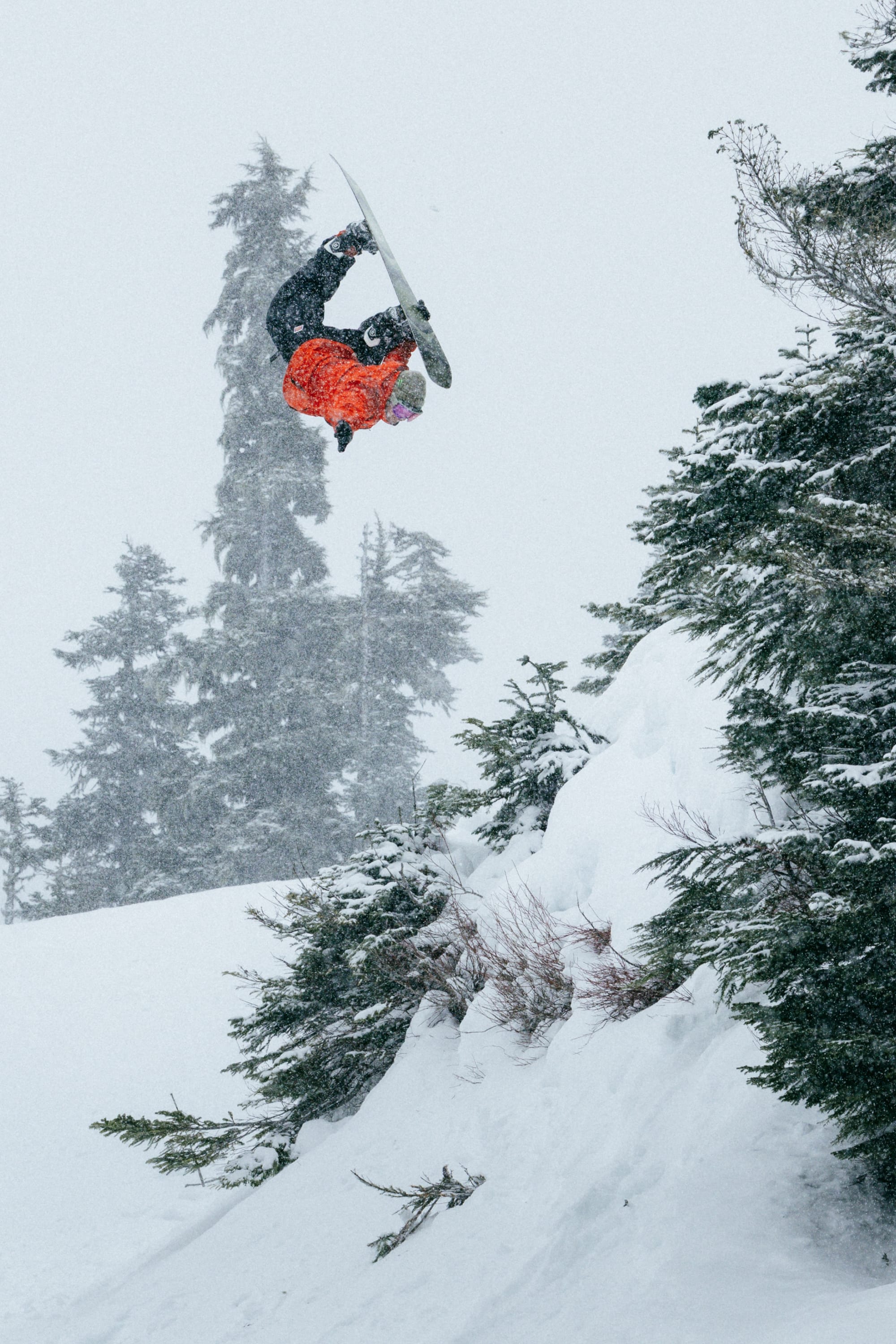 Wearing the 686 Men’s GORE-TEX® Core Shell Jacket, a snowboarder launches into a mid-air flip off a snowy slope, surrounded by snow-covered trees and falling snow.