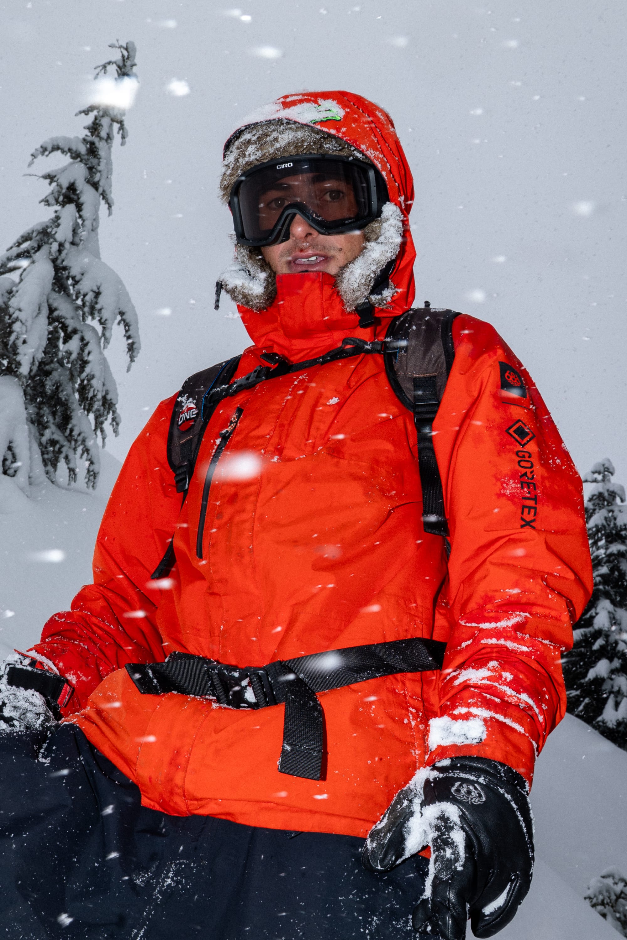 Wearing the 686 Men’s GORE-TEX® Core Shell Jacket, a person stands outdoors in falling snow, surrounded by snow-covered trees, with black gloves and snow goggles.