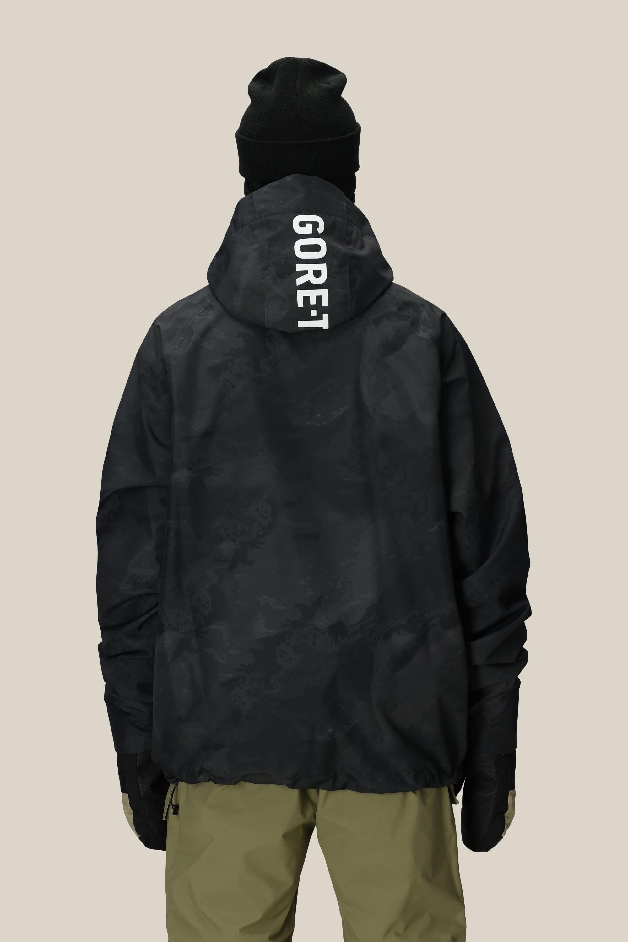 A person in a black beanie, black gloves, olive green pants, and a 686 Mens GORE-TEX Fragment Shell Anorak stands with their back to the camera against a plain background.