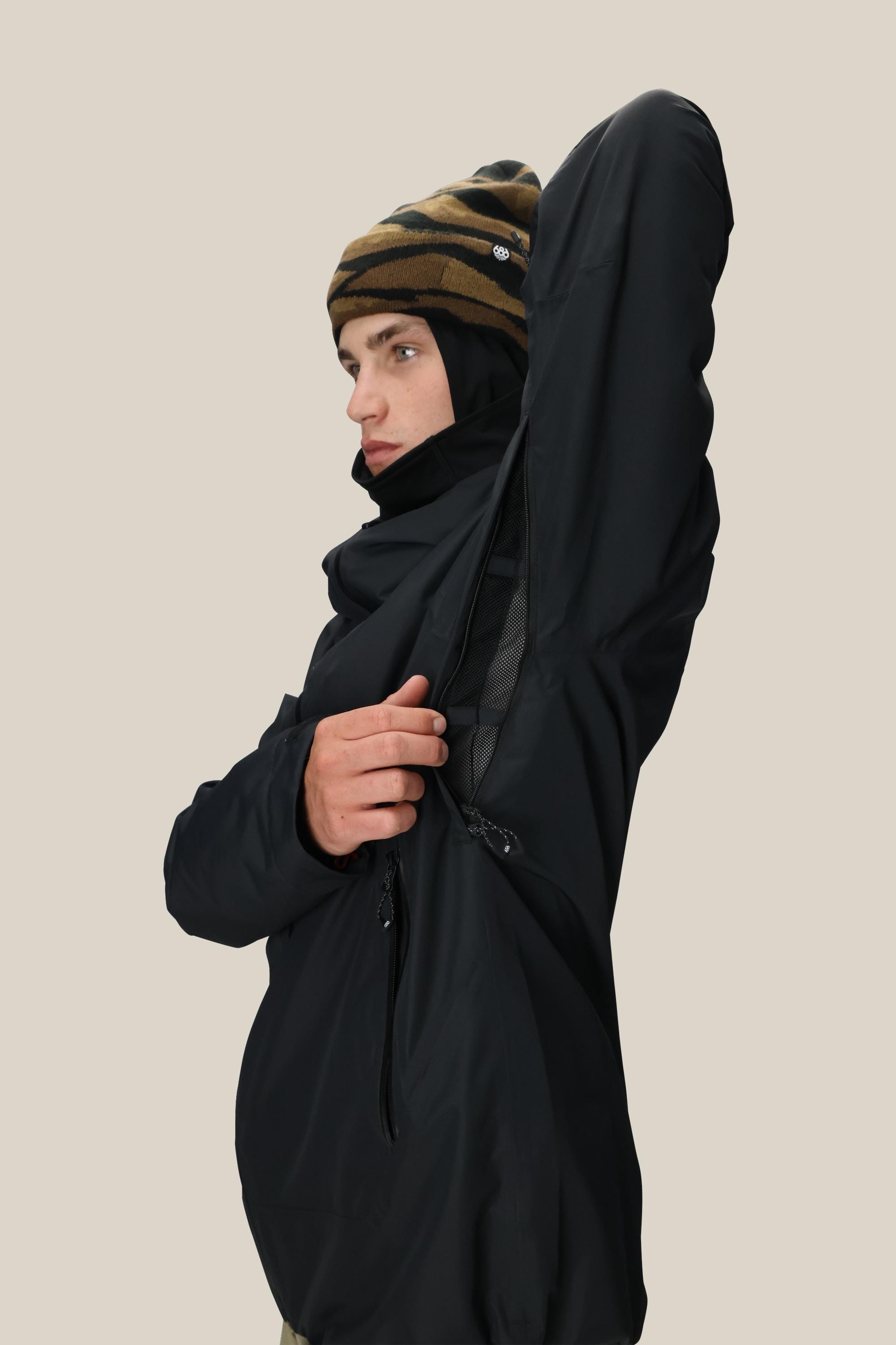 Wearing a 686 Mens Gateway Shell Jacket, a person opens a zippered underarm vent, complemented by a brown-striped beanie, set against a neutral beige background.