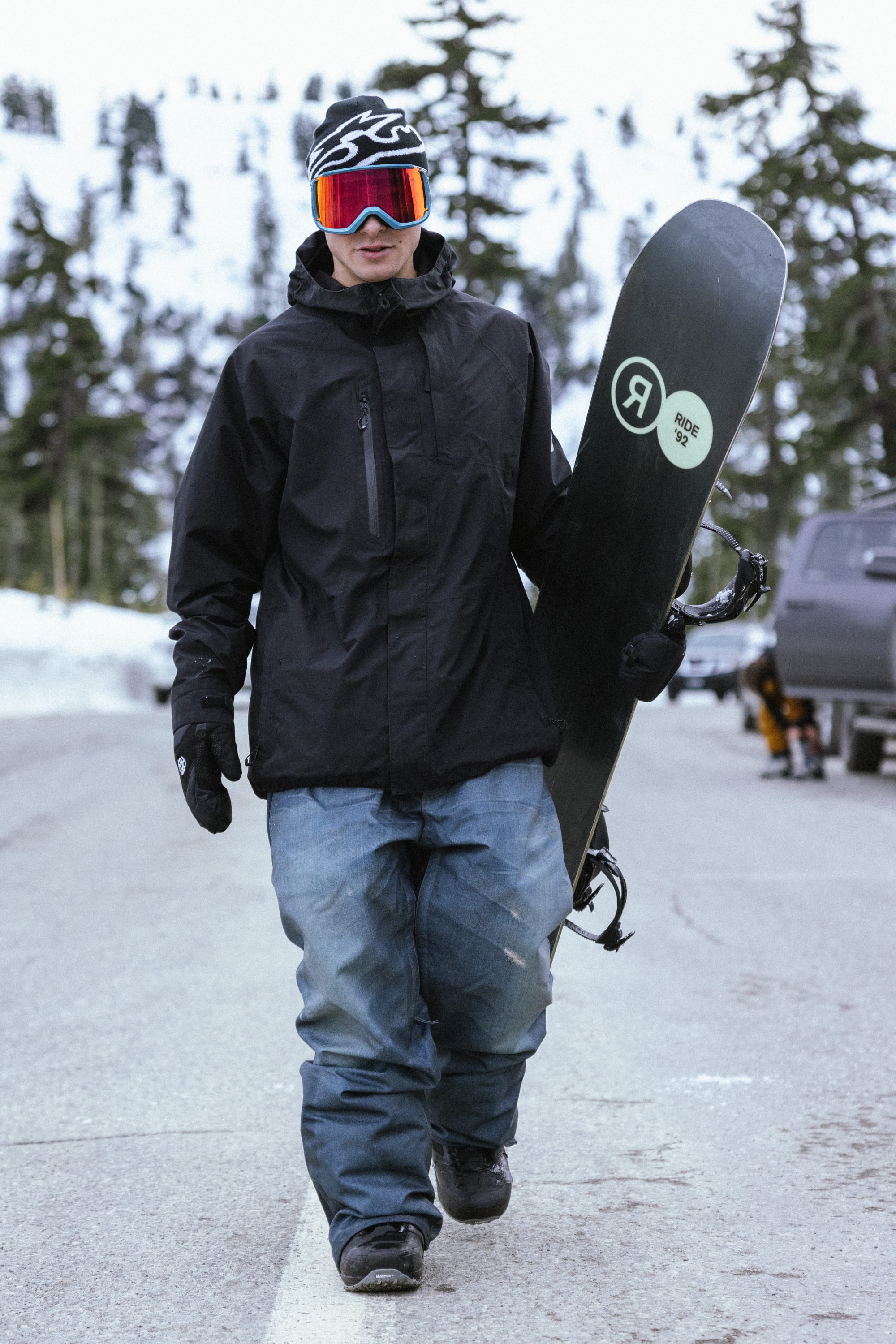 A man wearing the 686 Men’s GORE-TEX® Core Insulated Jacket walks outdoors on a snowy day, holding a snowboard upright, with cars and snow-covered trees visible in the background.