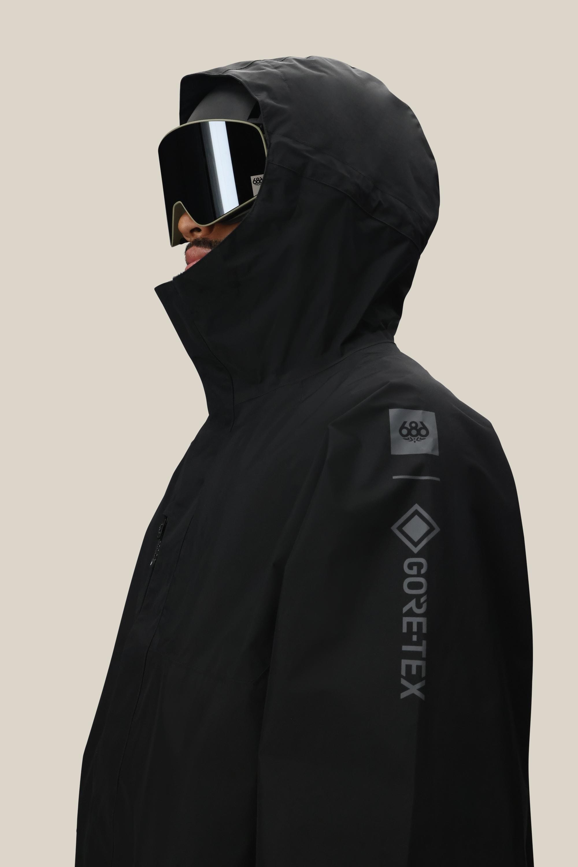 A person wearing the 686 Men’s GORE-TEX® Core Insulated Jacket in black, paired with large black ski goggles, is shown in profile against a plain beige background. The 686 and GORE-TEX® logos are visible on the sleeve.