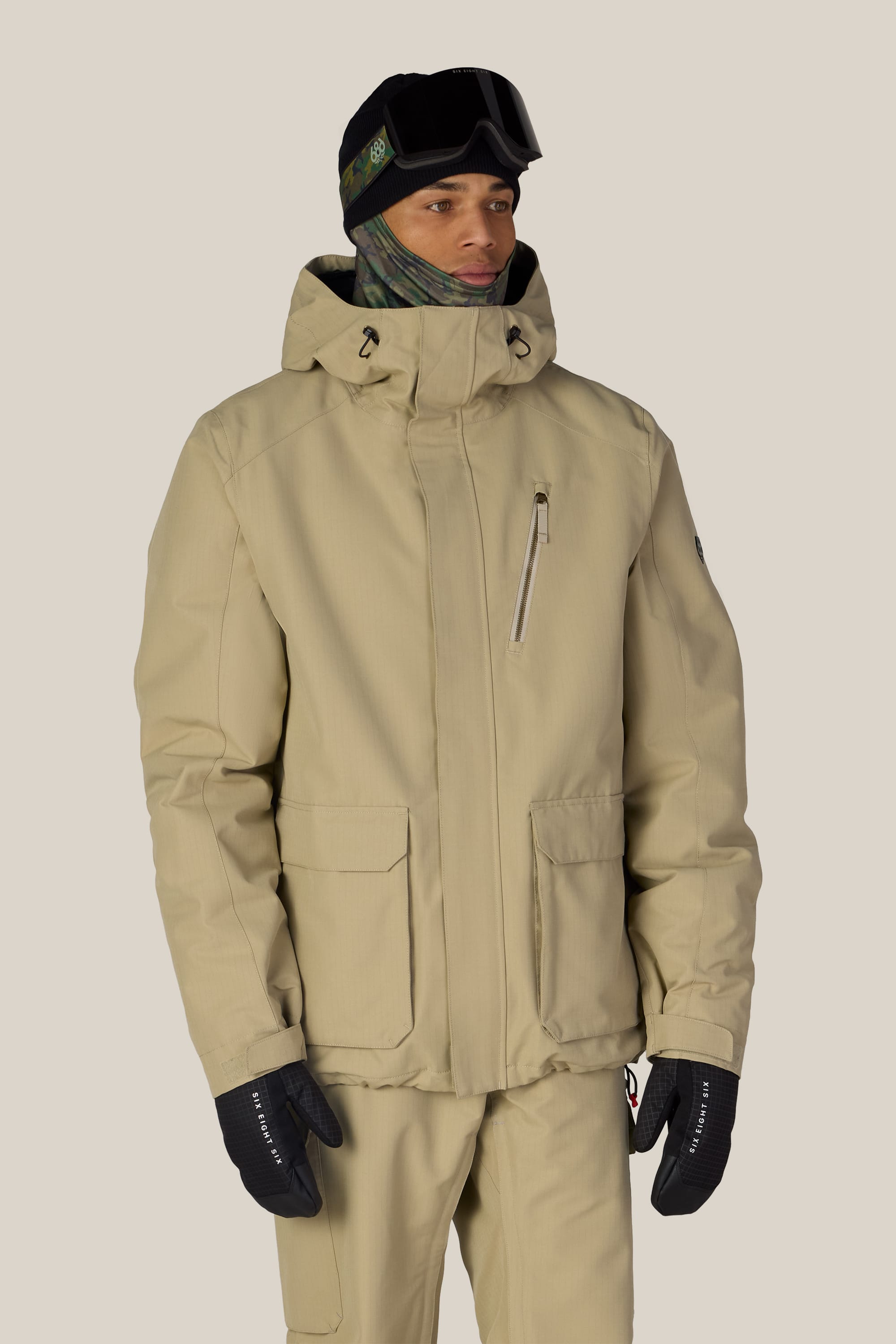 A person wearing the 686 Men’s Nightline™ Insulated Jacket, paired with matching pants, black gloves, a patterned balaclava, and black ski goggles stands against a plain light background.