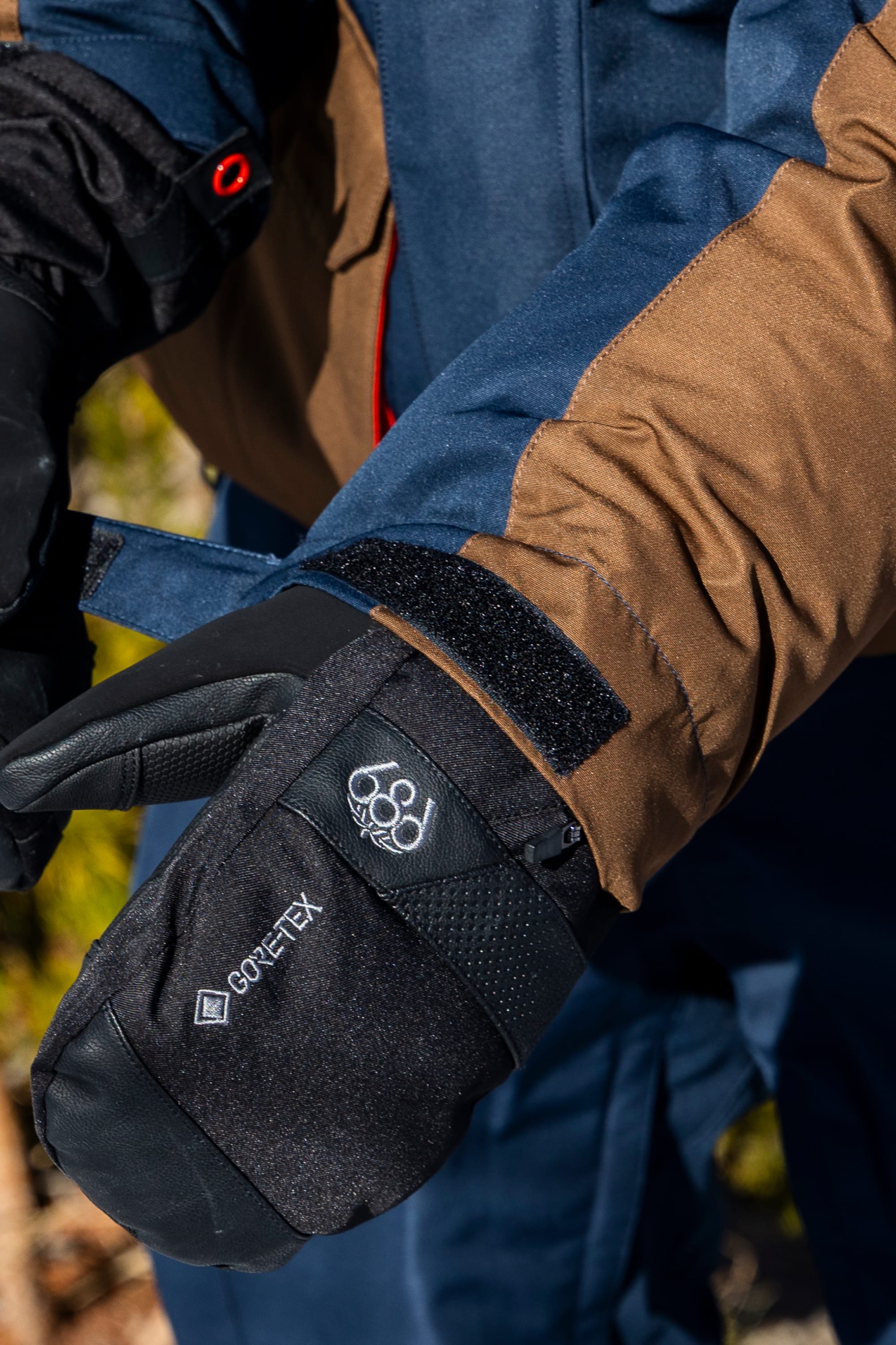 A person adjusts a velcro strap on their black Gore-Tex glove while wearing the 686 Men’s Nightline™ Insulated Jacket in blue and brown. The image focuses on the gloved hands and the sleeve of the 686 jacket, suggesting cold outdoor conditions.