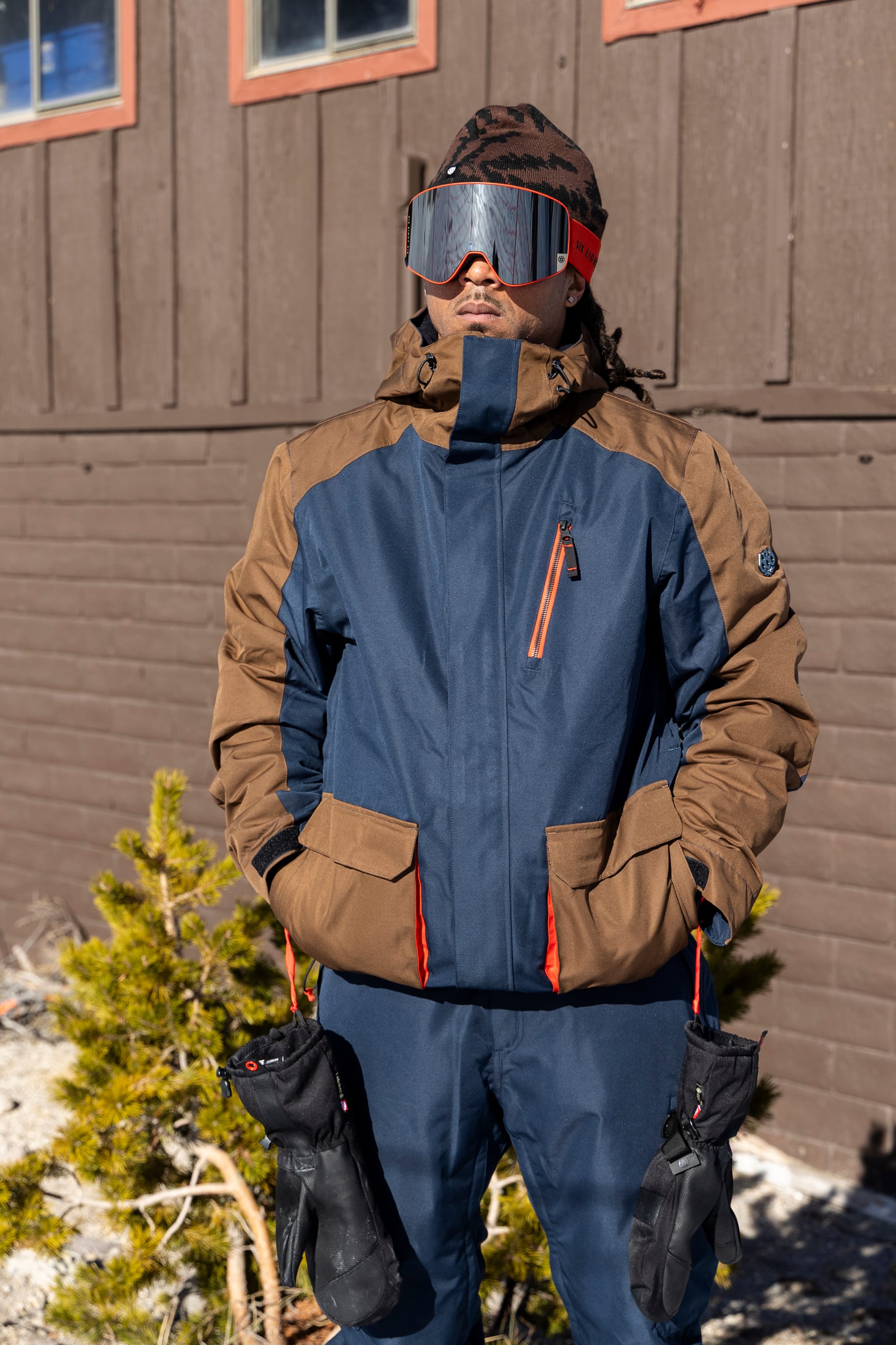 A person wearing the 686 Men’s Nightline™ Insulated Jacket and ski pants, along with reflective goggles, stands outside a wooden building holding gloves in each hand. Nearby, there is a small pine shrub and snow on the ground.