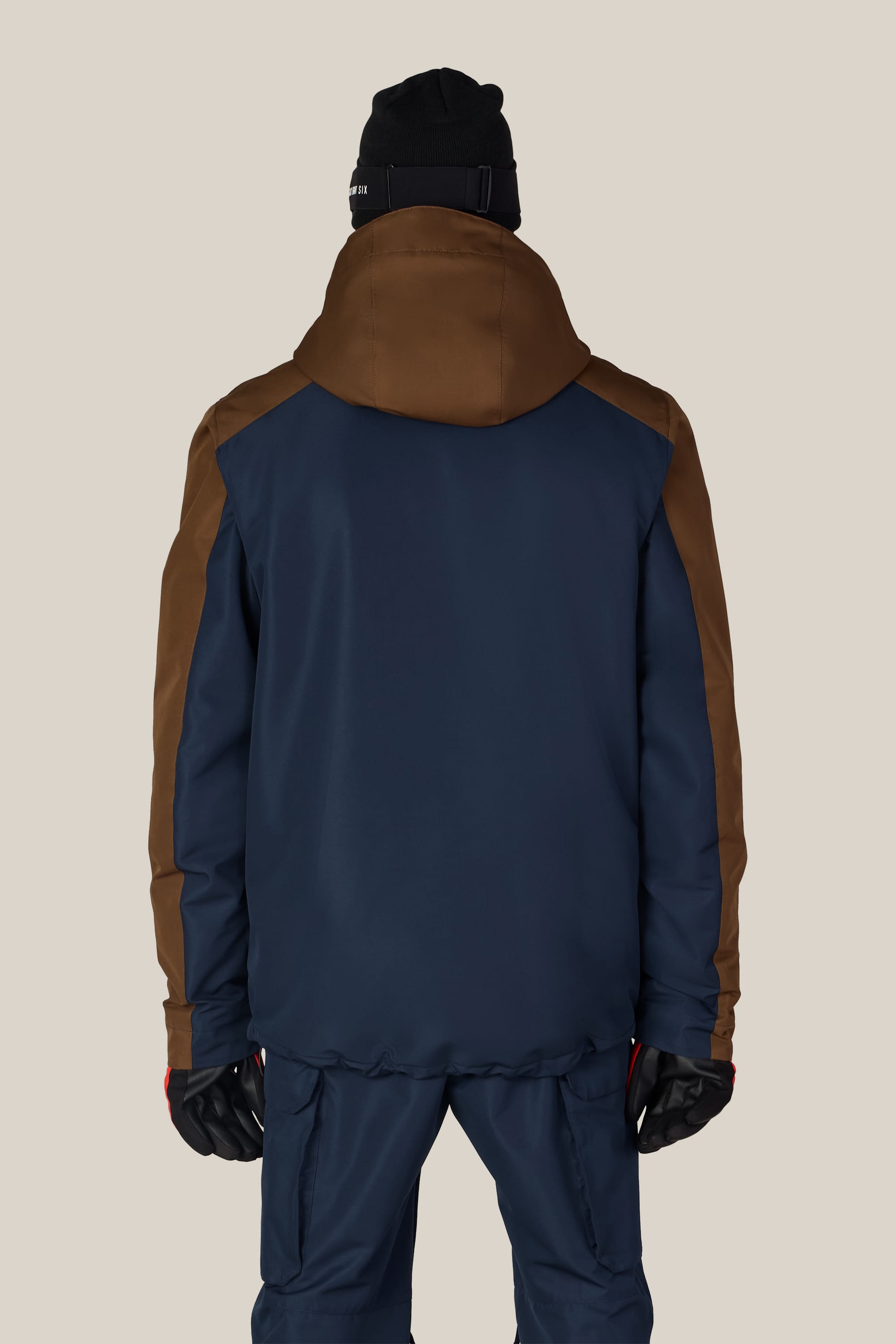 A person wearing the 686 Men’s Nightline™ Insulated Jacket by 686, styled in navy blue and brown, along with navy pants, black gloves, and a black beanie is viewed from the back against a plain beige background.