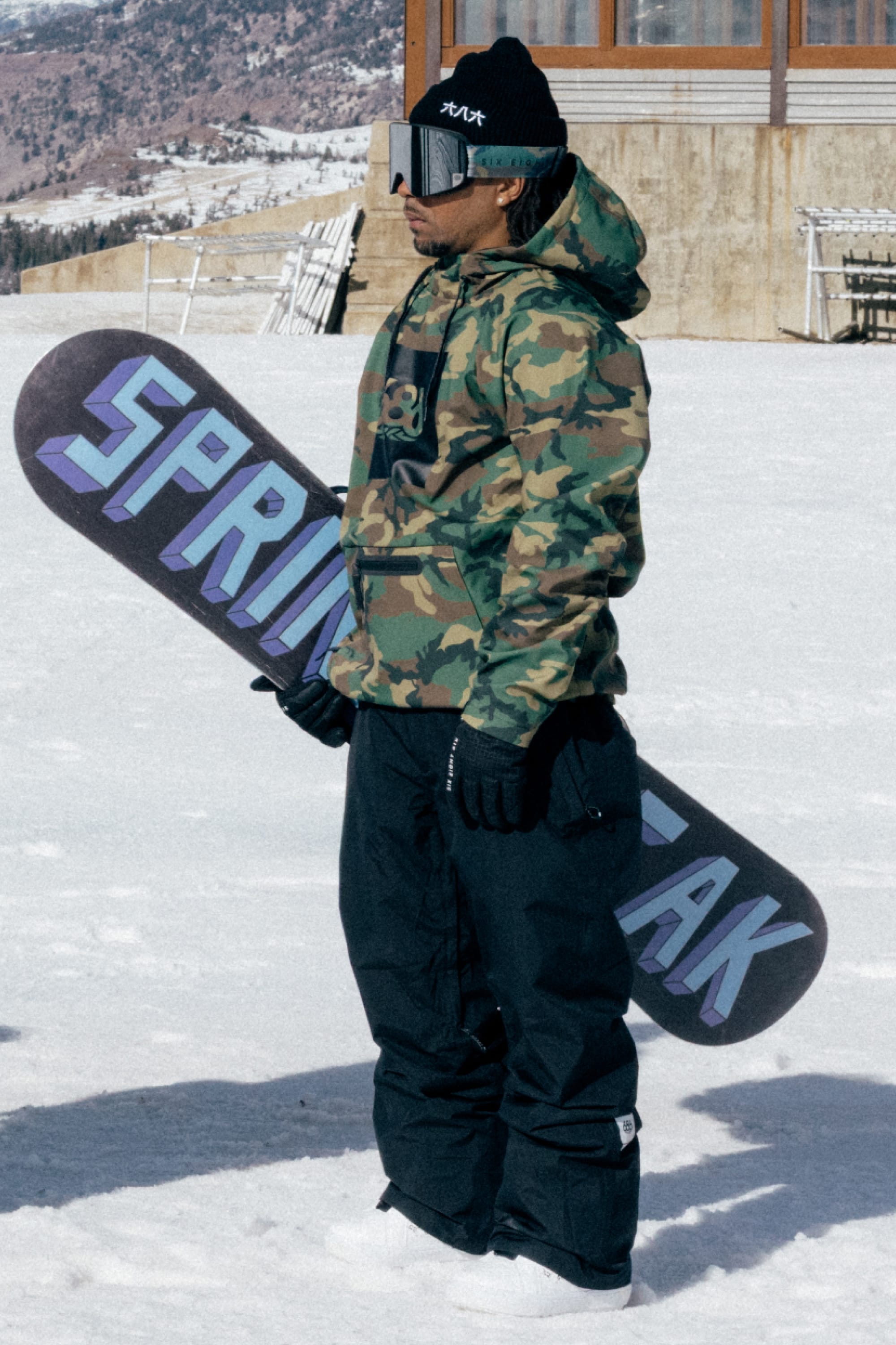 A person wearing the 686 Men’s GORE-TEX® Core Shell Pant, along with a camo jacket, gloves, and a beanie, stands on snow holding a snowboard that reads SPRING BREAK. Snowy mountains and a building are visible in the background.