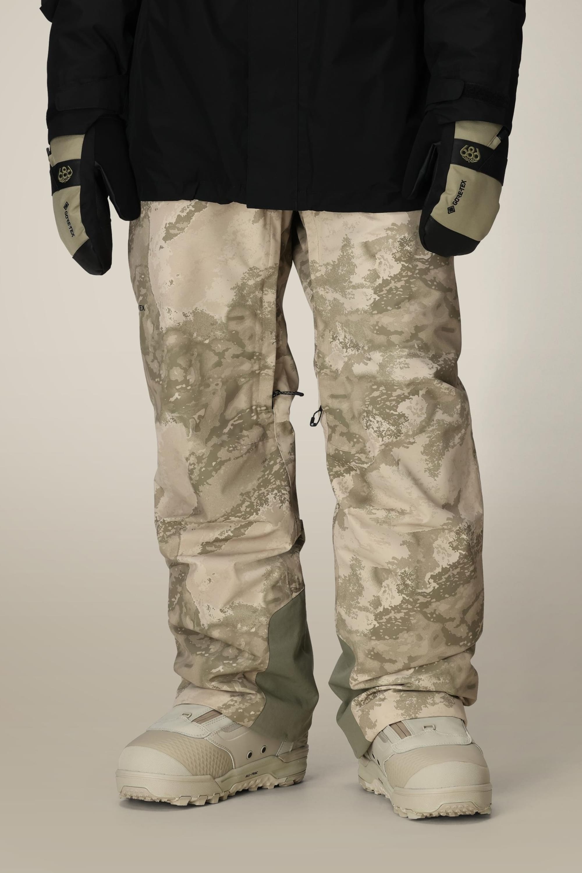 A person wears 686 Mens GORE-TEX Core Shell Pant in light beige and gray, with white snow boots, a black jacket, and black-and-tan gloves, standing indoors against a neutral background.