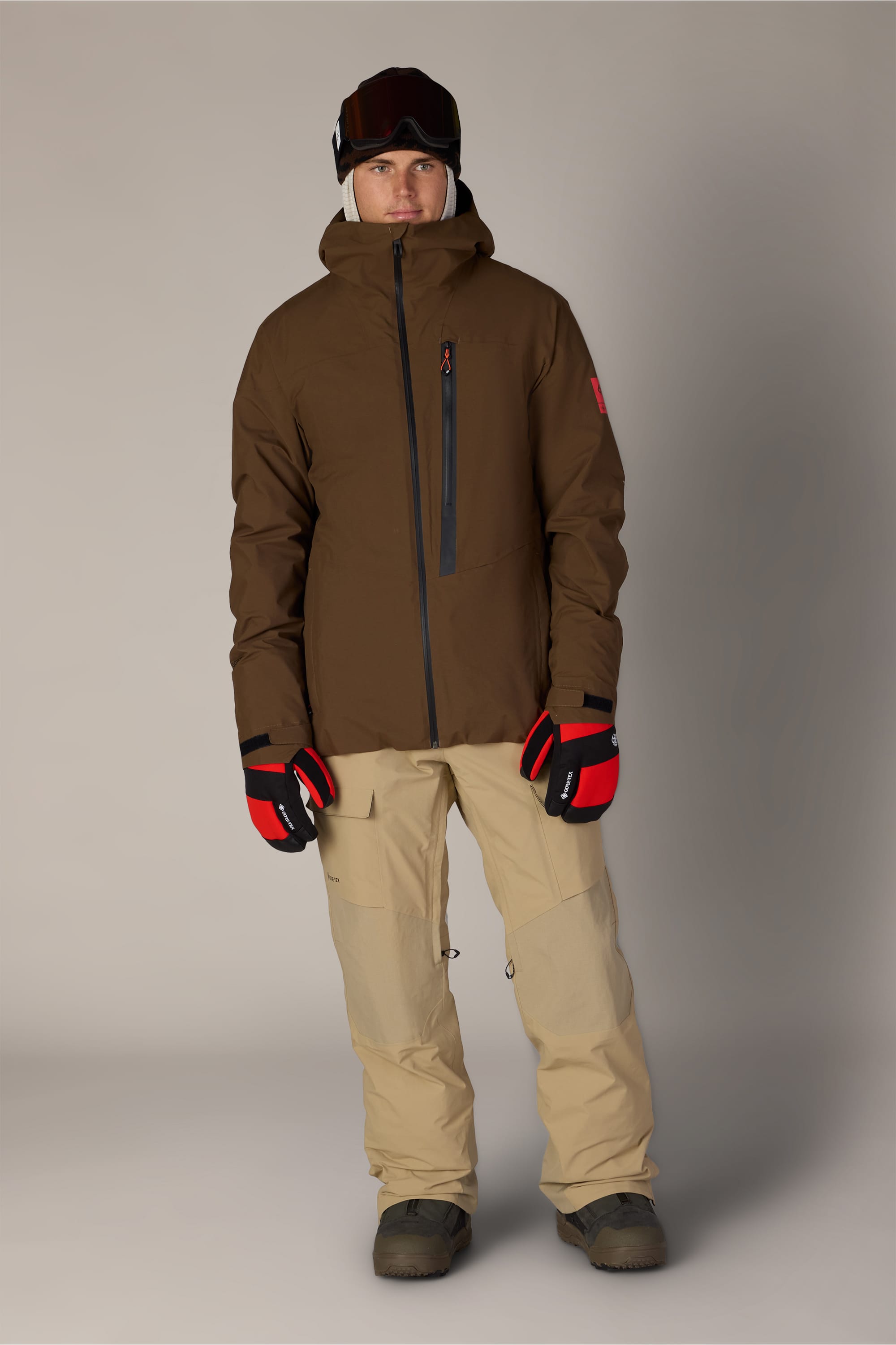 A person wearing a brown hooded jacket, red and black gloves, ski goggles, dark boots, and 686 Men’s GORE-TEX® Dispatch™ Shell Pant by 686 stands against a plain gray background.