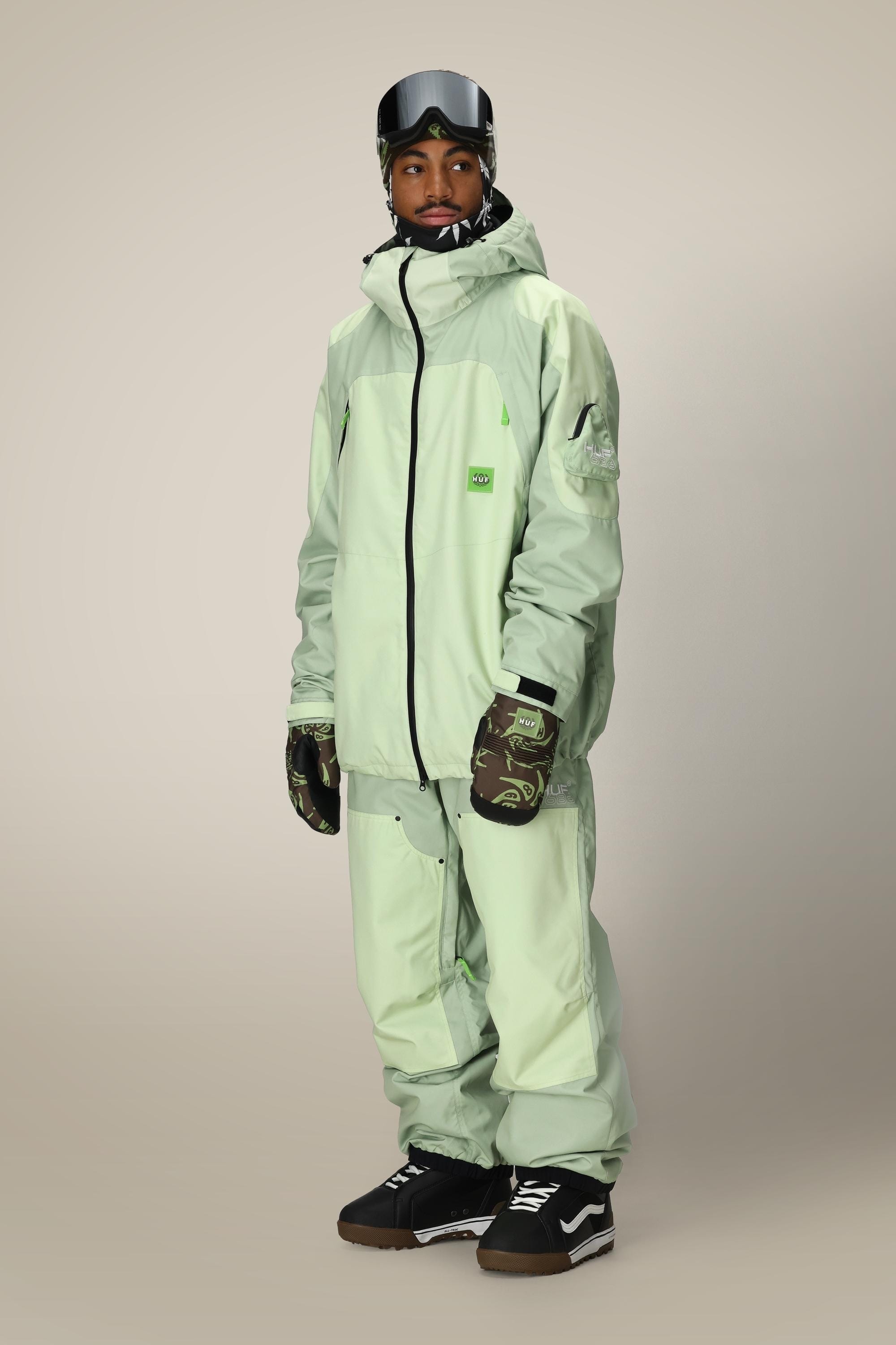 A person poses against a beige background wearing a mint green ski jacket, patterned gloves, black snow boots, a printed neck gaiter, a dark ski visor, and 686 Mens HUF Double H Shell Pant by 686.