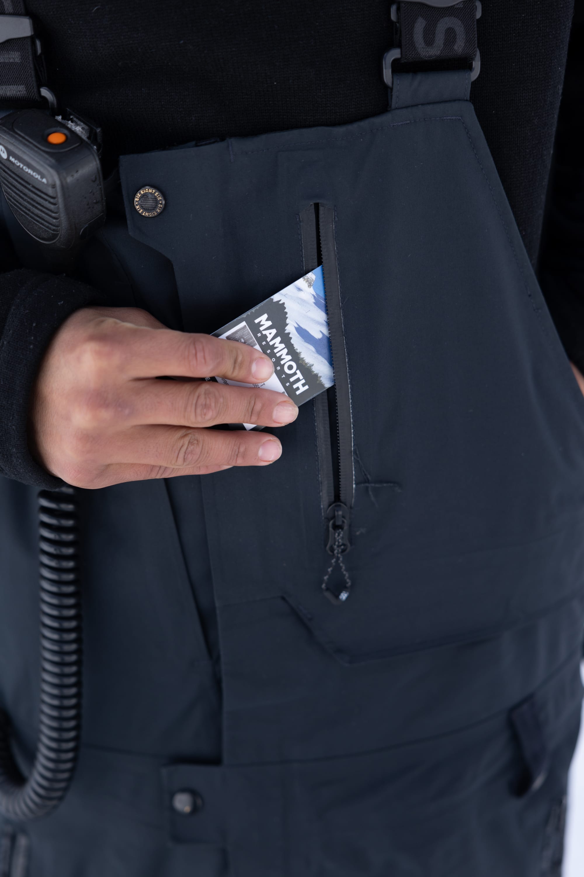 Outfitted in the 686 Men’s GORE-TEX® Dispatch™ Shell Bib, a person slips a Mammoth Mountain pass into a zippered chest pocket while a two-way radio is clipped to their shoulder.