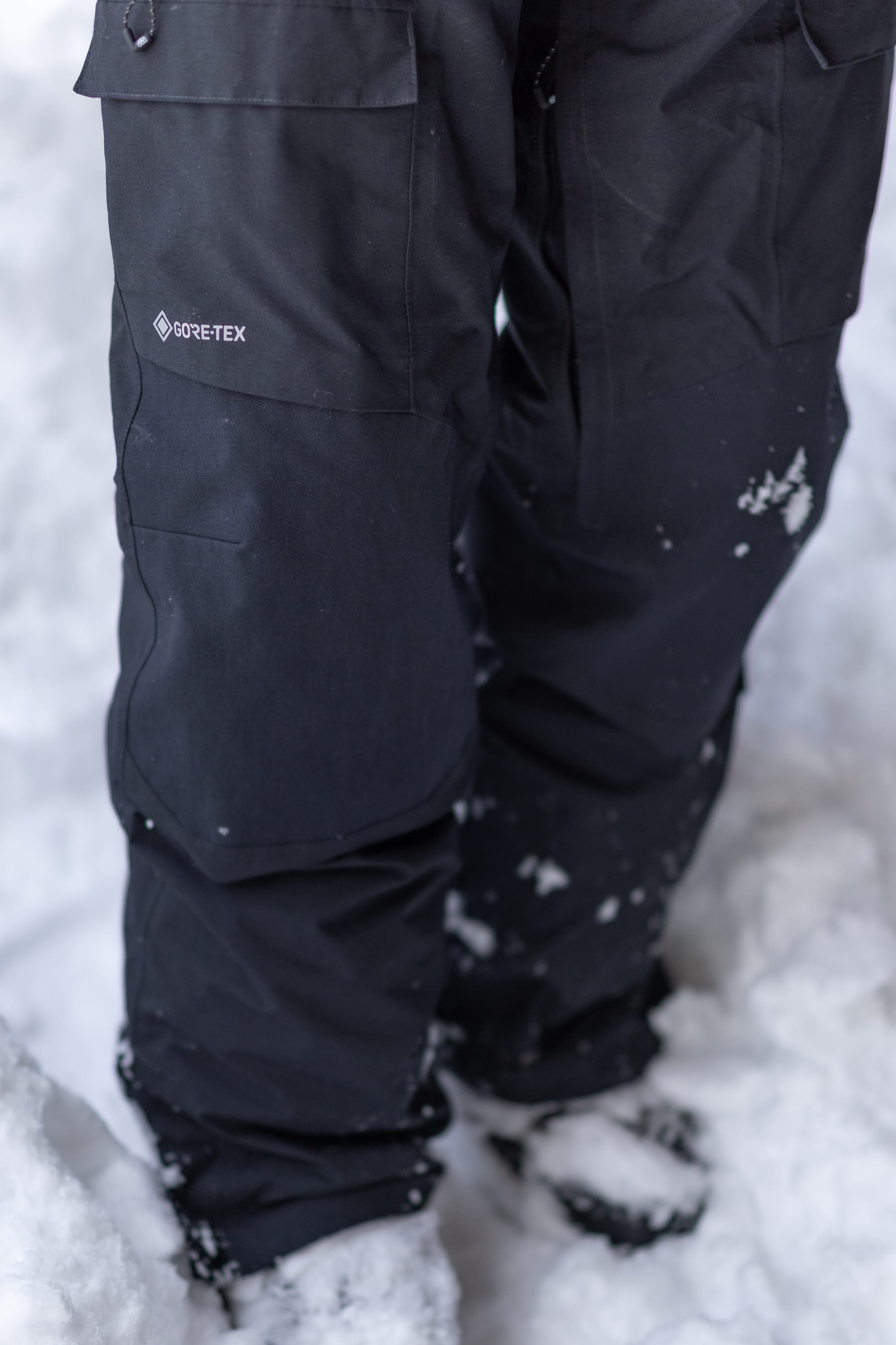 A person wearing 686 Men’s GORE-TEX® Dispatch™ Shell Bib snow pants stands in the snow, with some snow clinging to the lower legs and boots.