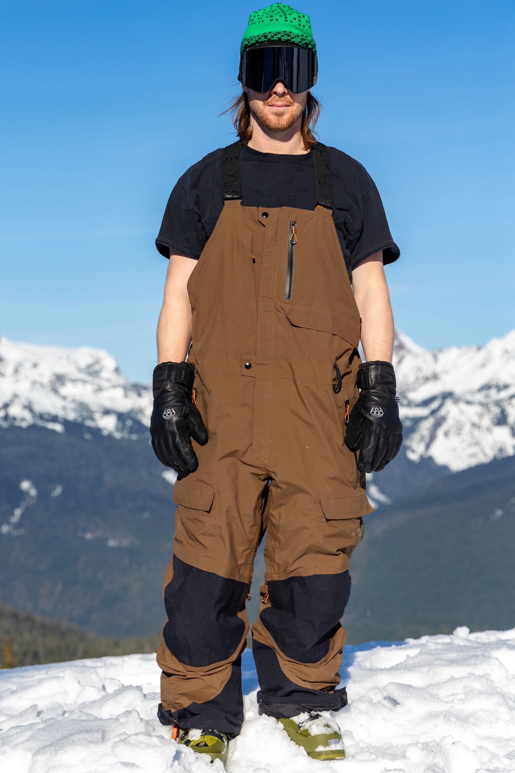 A person wearing the 686 Men’s GORE-TEX® Dispatch™ Shell Bib by 686, along with a black shirt, black gloves, green hat, and ski goggles, stands on snowy ground with mountains and a blue sky in the background.