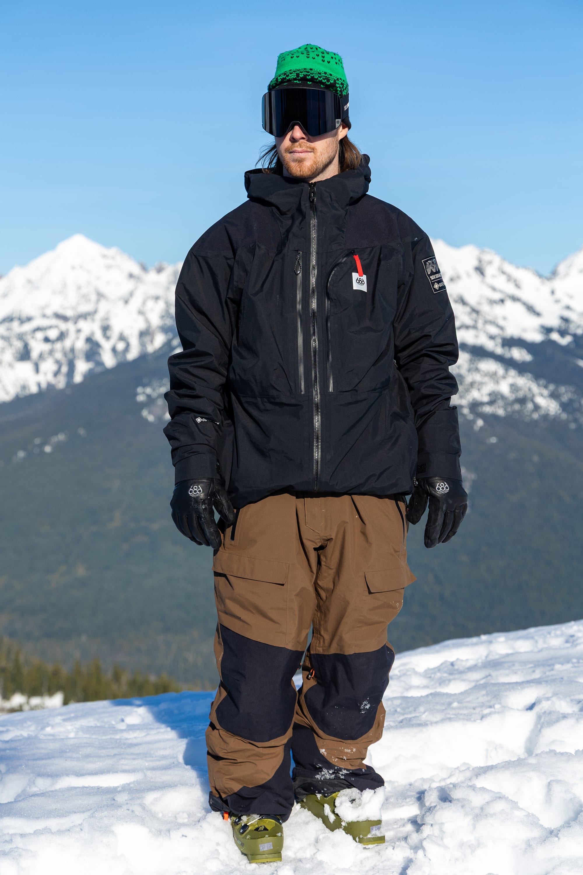 Wearing the 686 Men’s GORE-TEX® Dispatch™ Shell Bib by 686, a person in a black jacket, green helmet, and ski goggles stands on snow with mountain peaks and a blue sky in the background.