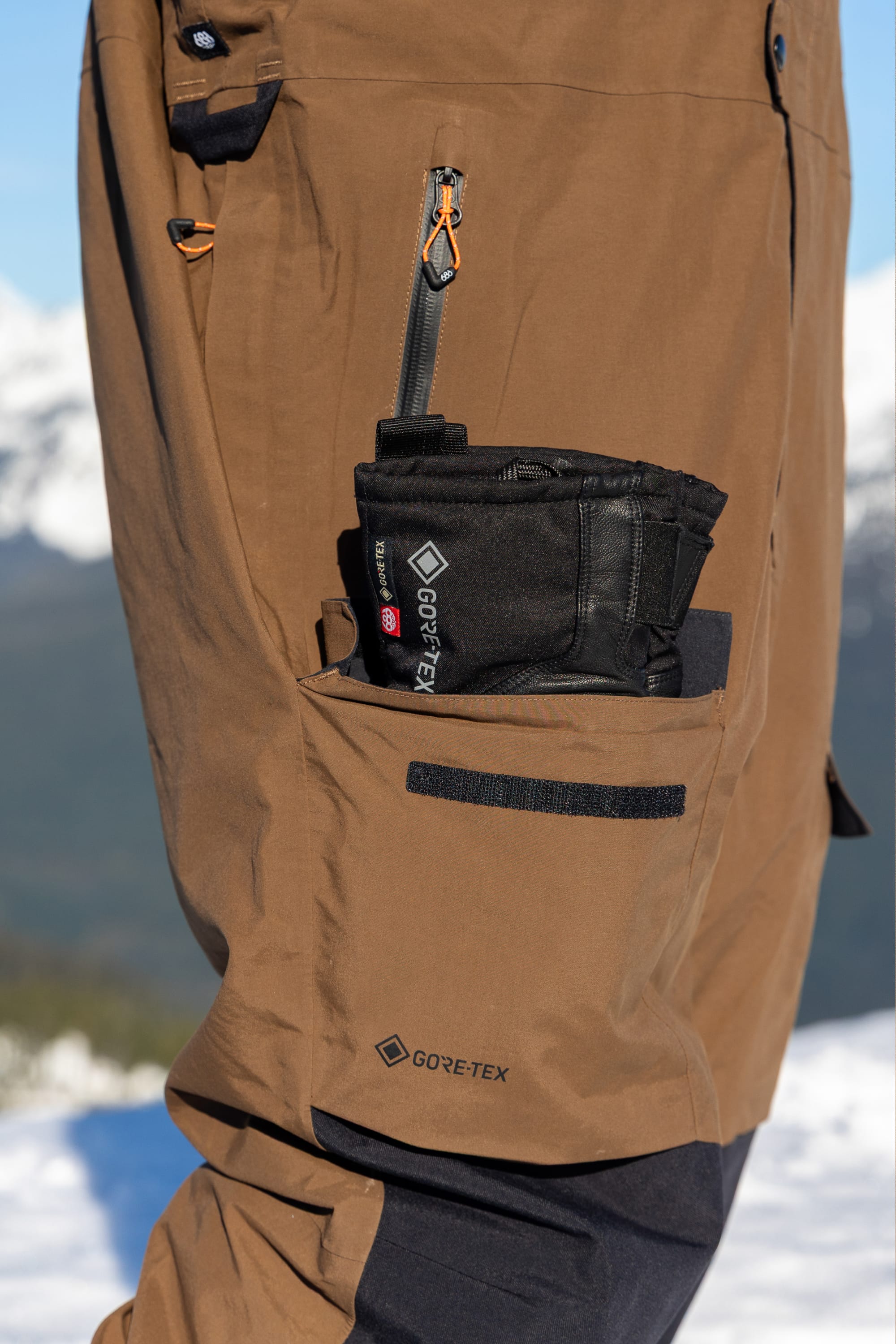 A close-up of the 686 Men’s GORE-TEX® Dispatch™ Shell Bib in brown, featuring a black pouch in the side pocket, photographed outdoors with snowy mountains and a blue sky in the background.