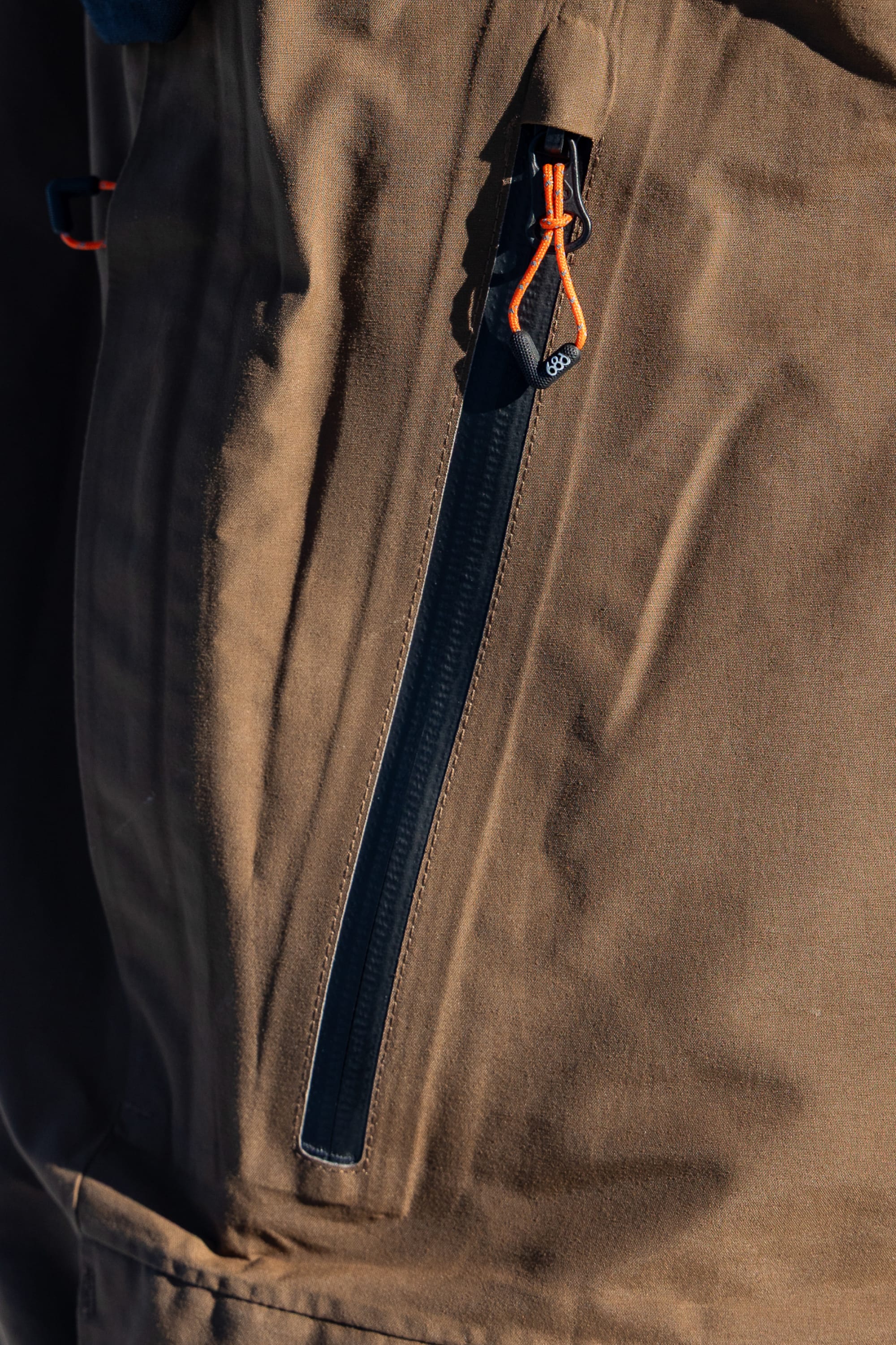 A close-up of the 686 Men’s GORE-TEX® Dispatch™ Shell Bib, featuring a brown fabric with a vertical zippered pocket, black zipper, and orange zipper pull.