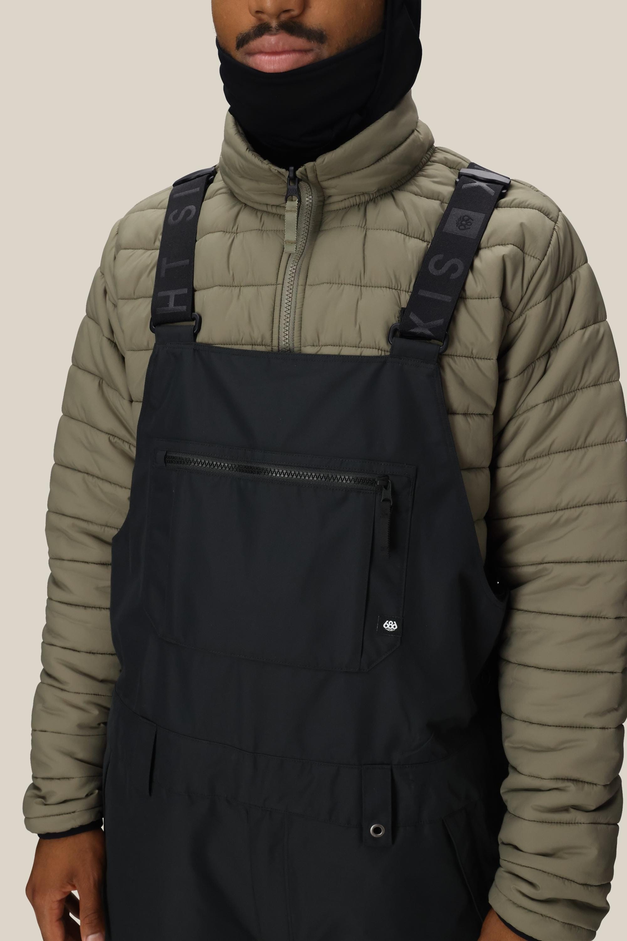 A person wears a light olive puffer jacket and 686 Mens 20K Hot Lap Shell Bib overalls in black, featuring a front zipper pocket and black straps. They have a black balaclava covering their neck and lower face against a plain beige background.