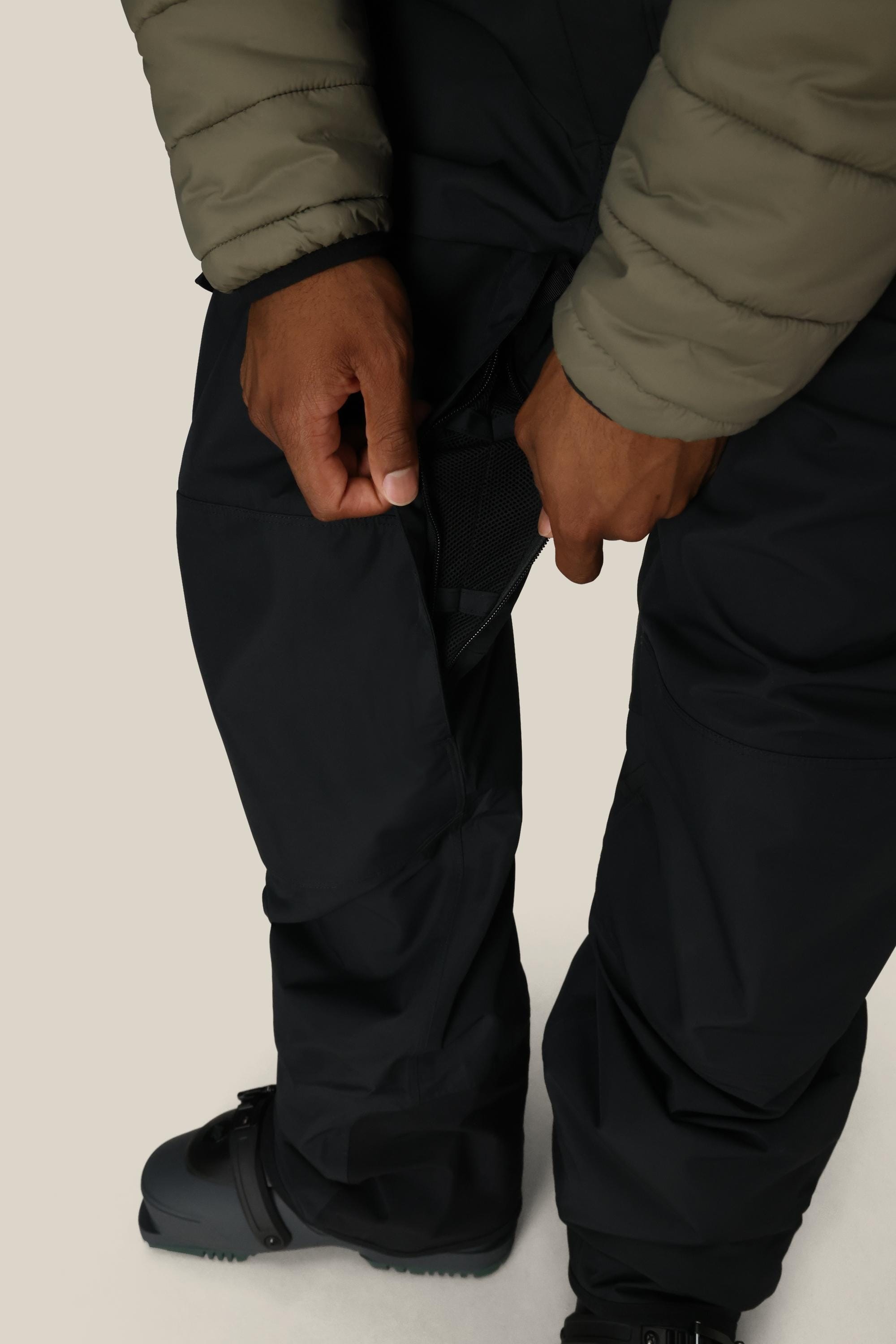 A person in a green jacket and 686 Mens 20K Hot Lap Shell Bib from 686 is unzipping a side vent, showing the mesh lining. Theyre also wearing black snow boots. The background is plain and light-colored.