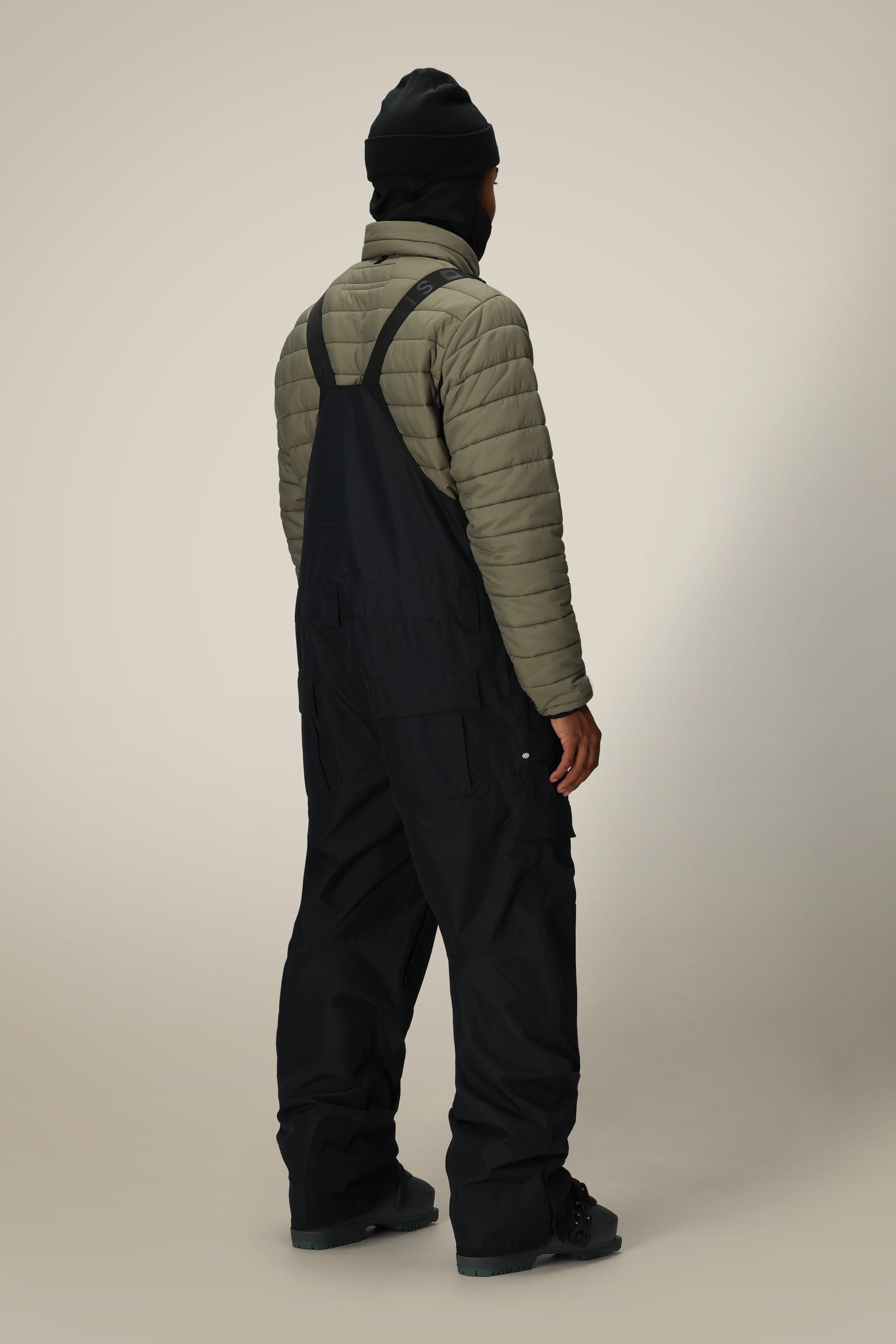 A person stands facing away in a black beanie, olive green quilted jacket, and the 686 Mens 20K Hot Lap Shell Bib by 686, made from polyester nylon spandex, paired with boots against a plain light background.