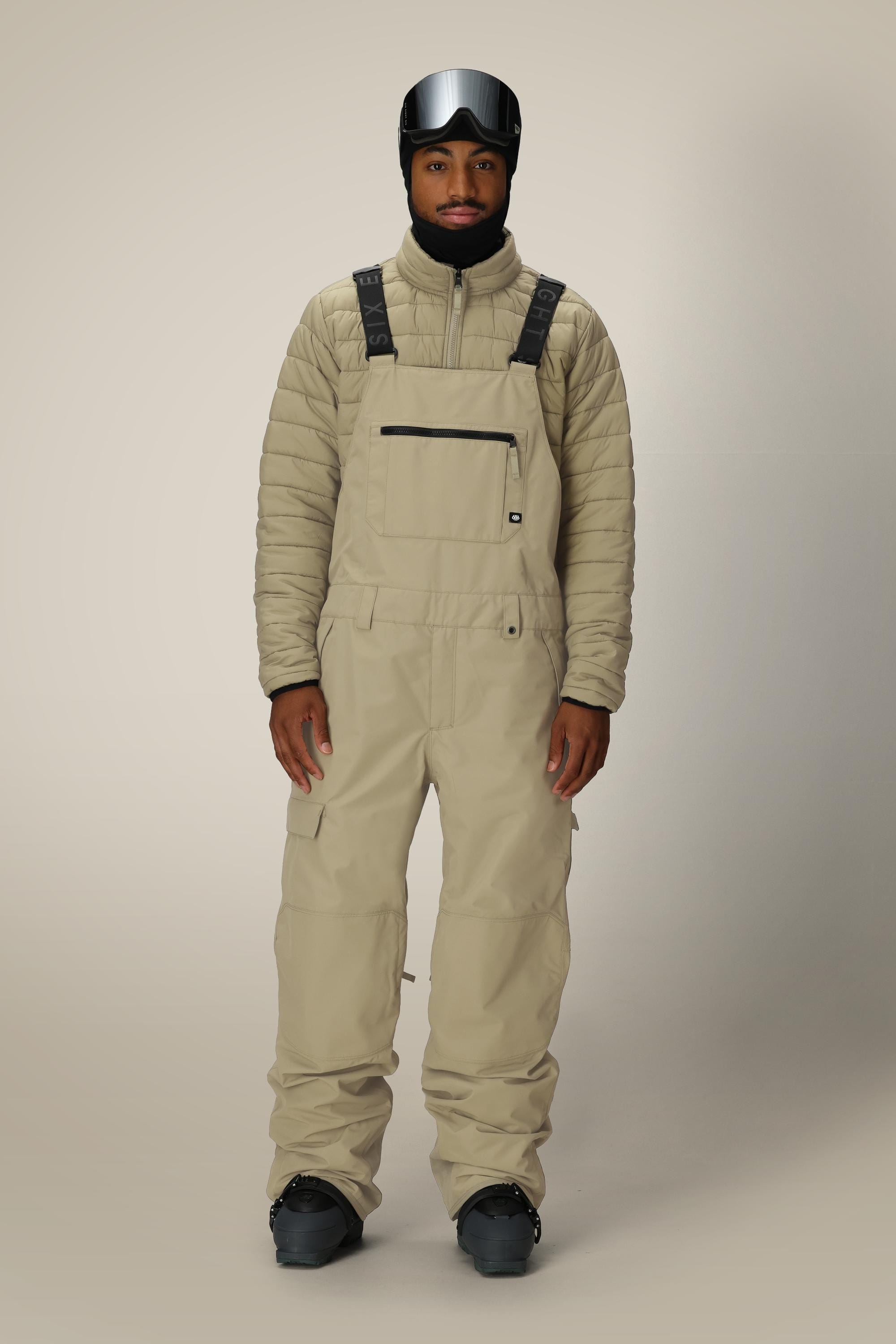 A man wearing the 686 Mens 20K Hot Lap Shell Bib, black boots, a puffy jacket, and a black ski helmet with goggles stands against a plain light background.