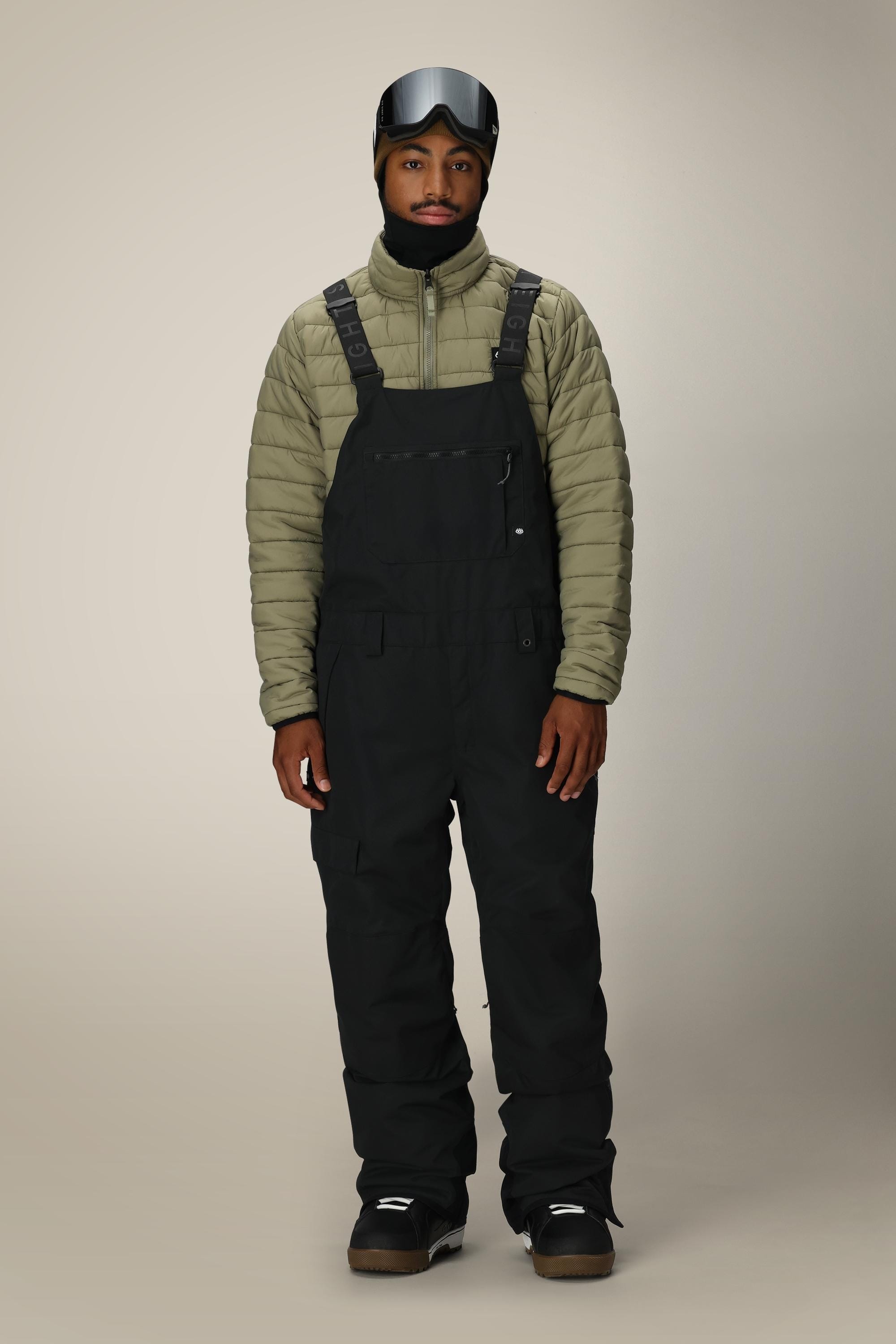 A person wearing a green puffer jacket, 686 Men’s Hot Lap™ Insulated Bib snow pants by 686, winter boots, and a ski mask with goggles stands facing forward against a plain beige background.