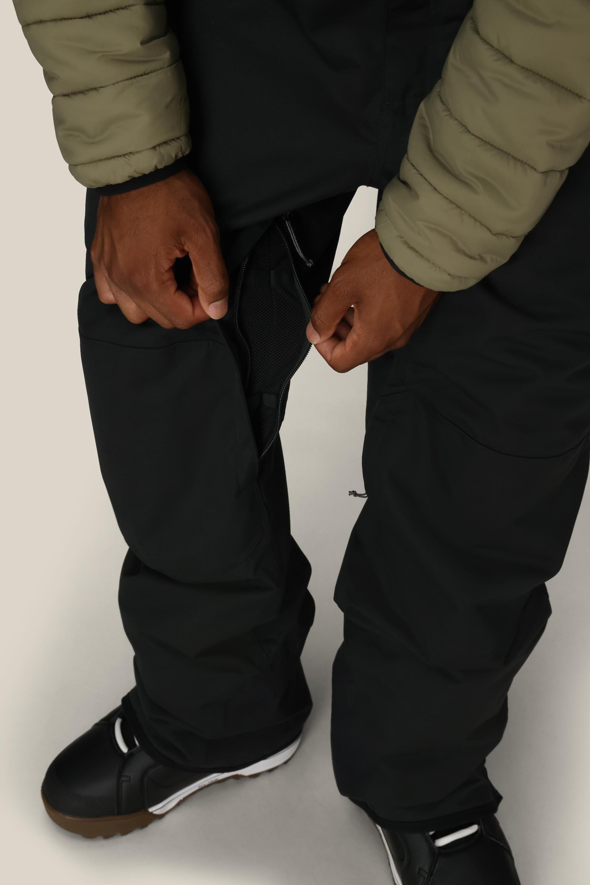 A person in a green jacket unzips a vent on the side of their black 686 Men’s Hot Lap™ Insulated Bib, revealing a mesh lining. They are wearing black snow boots.