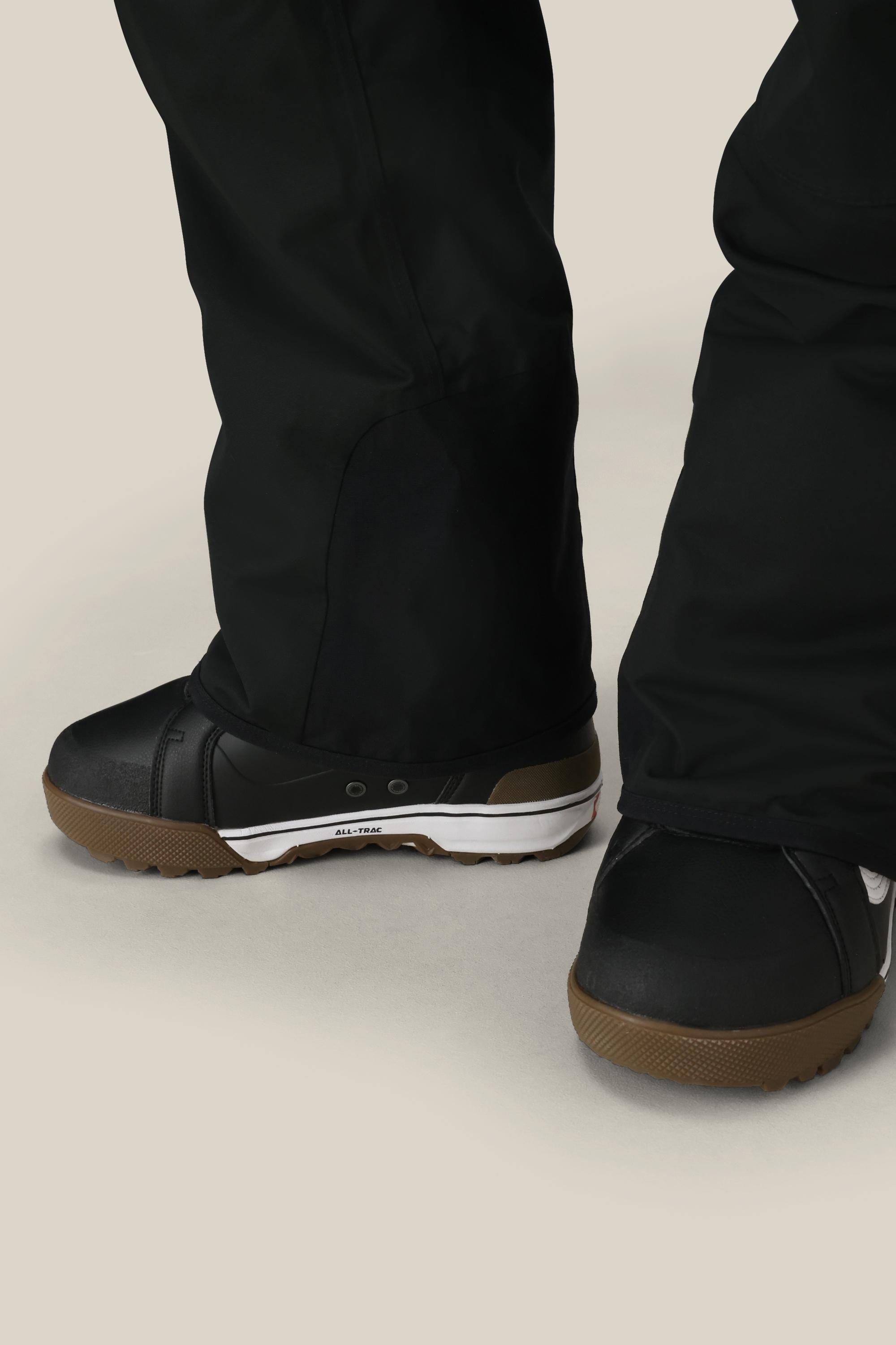 A person wearing the 686 Men’s Hot Lap™ Insulated Bib by 686 and black snow boots with brown and white soles, standing on a light-colored surface.