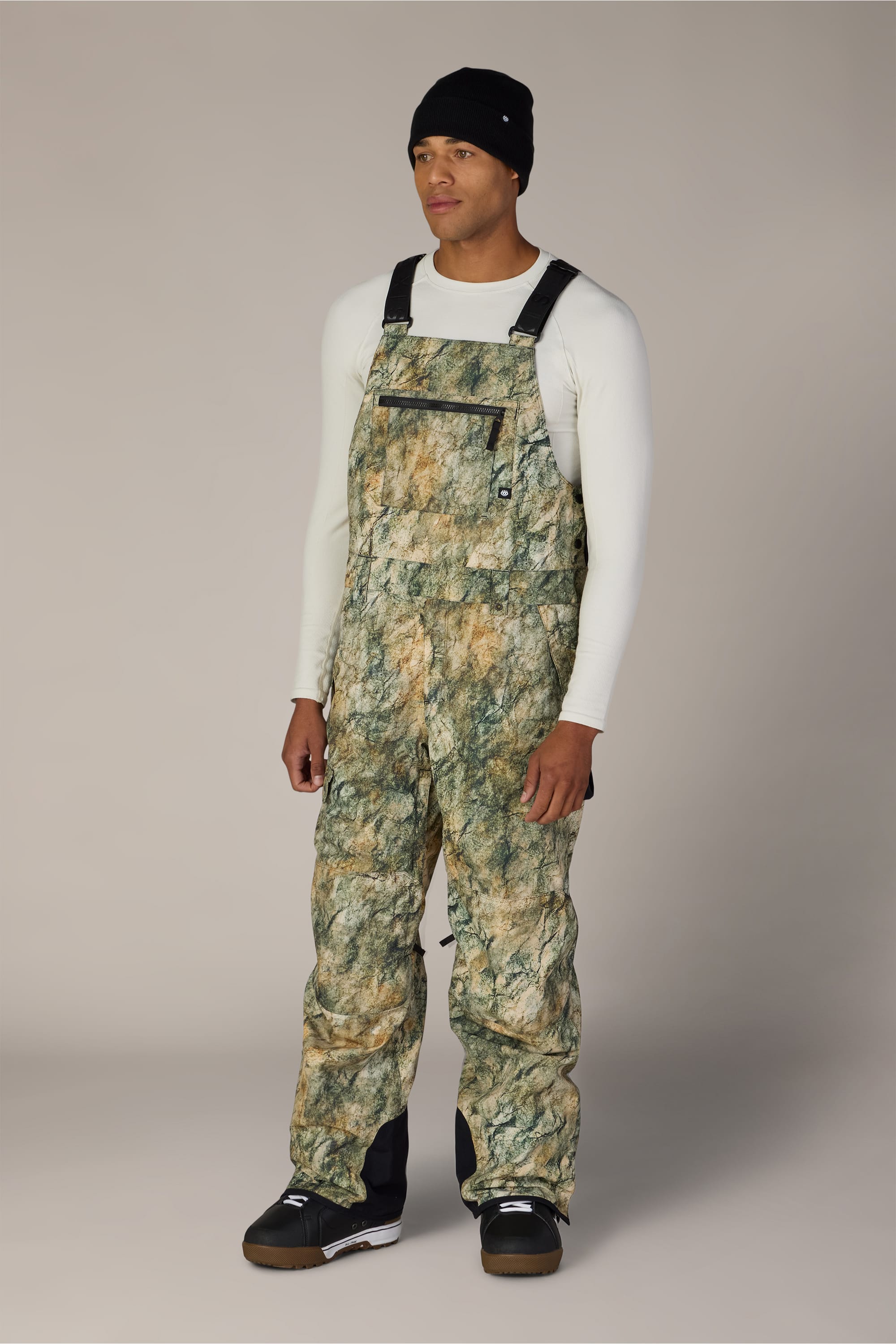 A person stands facing forward against a plain light gray background, wearing the 686 Men’s Hot Lap™ Insulated Bib by 686 in a camo pattern, paired with a white long-sleeve shirt and black beanie, with hands relaxed at their sides.
