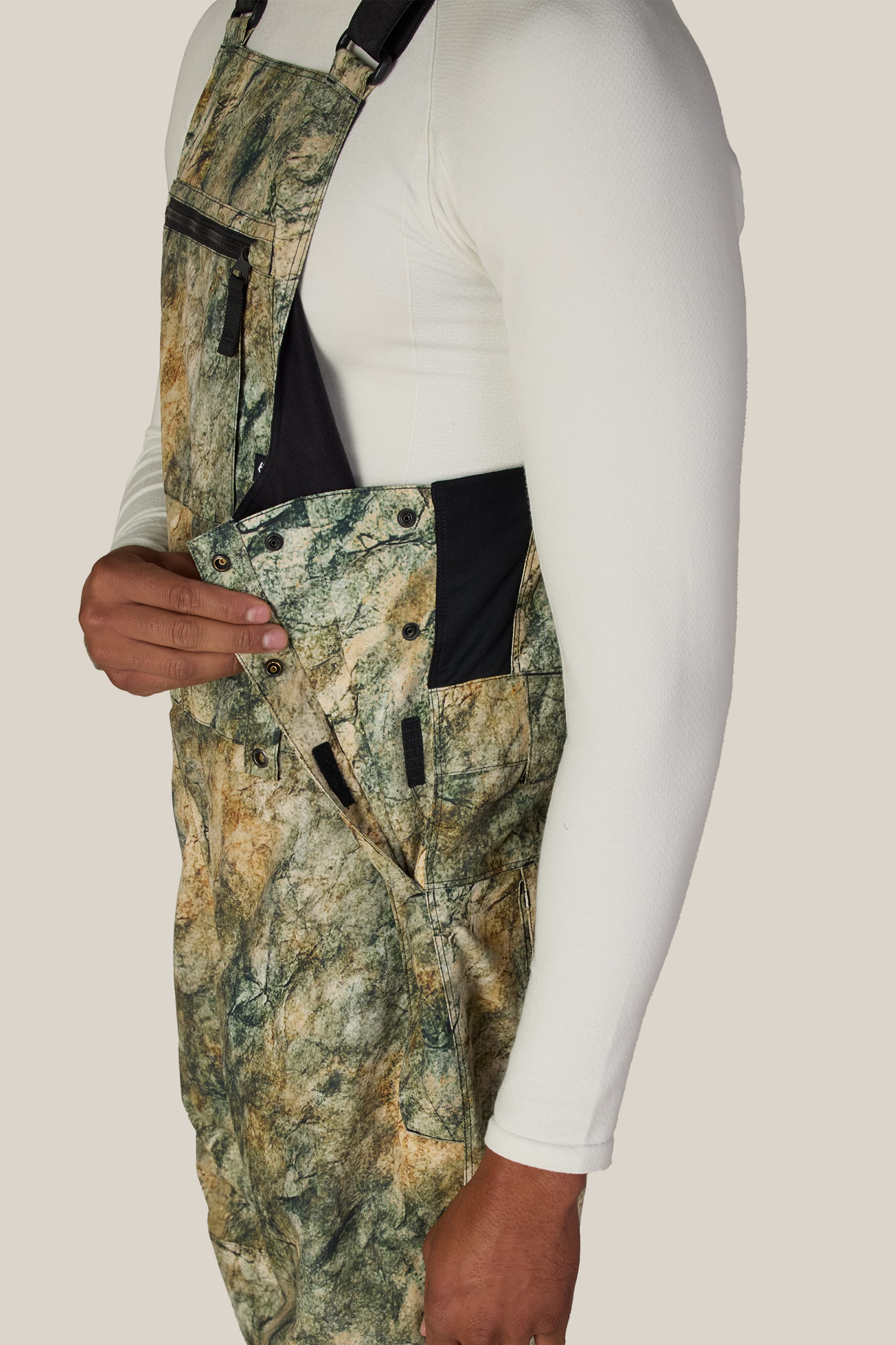 A person wearing the 686 Men’s Hot Lap™ Insulated Bib in a camouflage pattern and a white long-sleeve shirt unbuttons the side closure, showcasing the details of the 686 garment against a plain background.