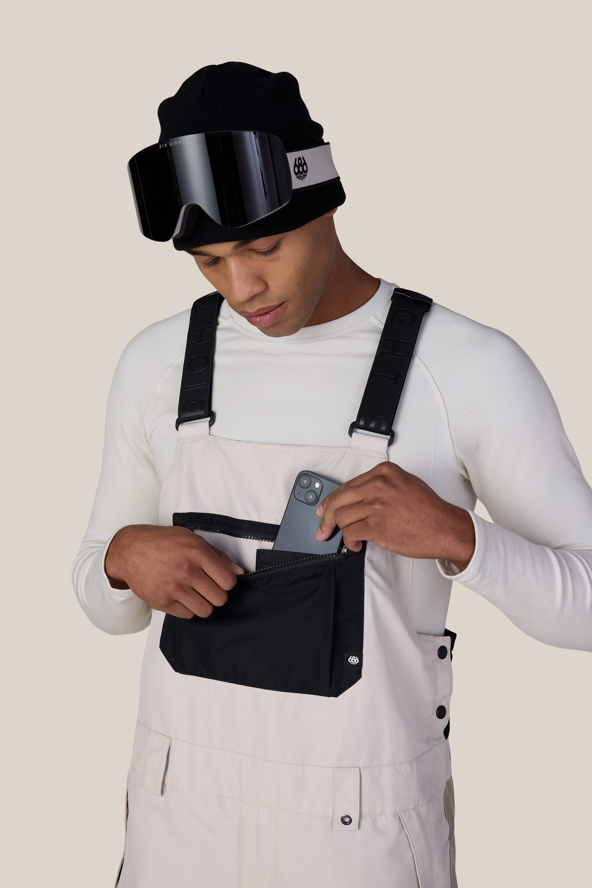 A person wearing a black beanie, ski goggles, and the 686 Men’s Hot Lap™ Insulated Bib by 686 in a light color places a smartphone into a black front pocket. The background is plain and beige.