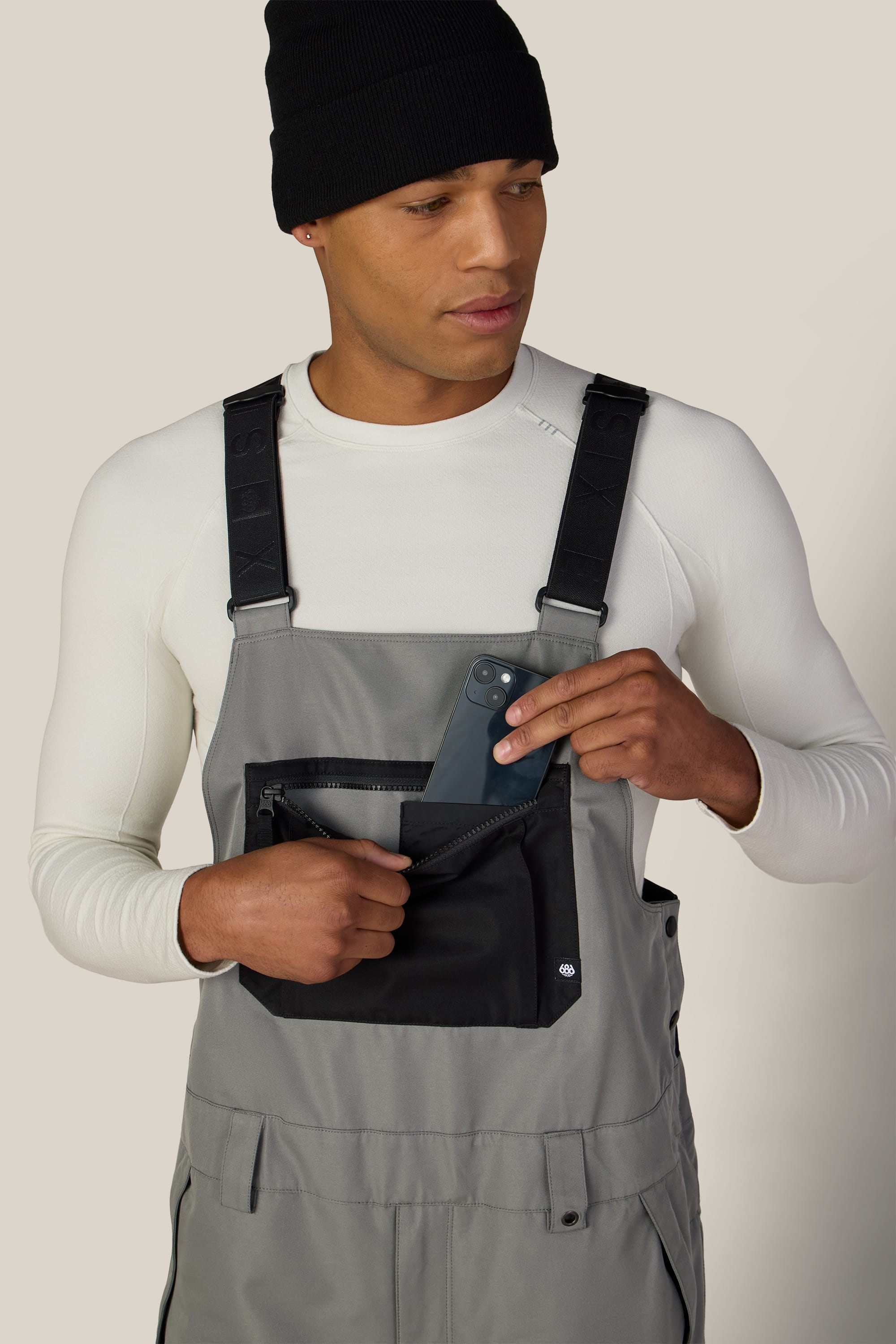 A person wearing the 686 Men’s Hot Lap™ Insulated Bib by 686, along with a black beanie and white shirt, places a smartphone into the zippered chest pocket of their overalls while looking down and to the side.