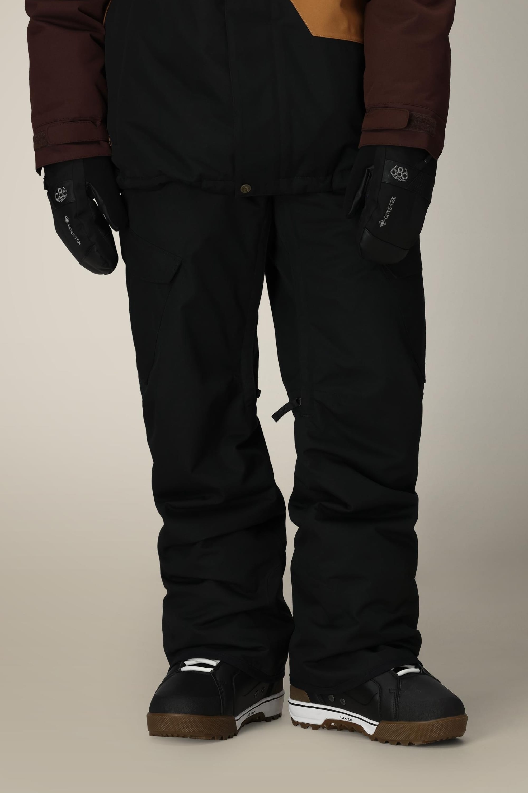A person wearing 686 Mens Infinity Insulated Cargo Pant stands indoors against a neutral background. Only the lower half of the body is visible, showing black snowboarding boots with white accents and matching black gloves.