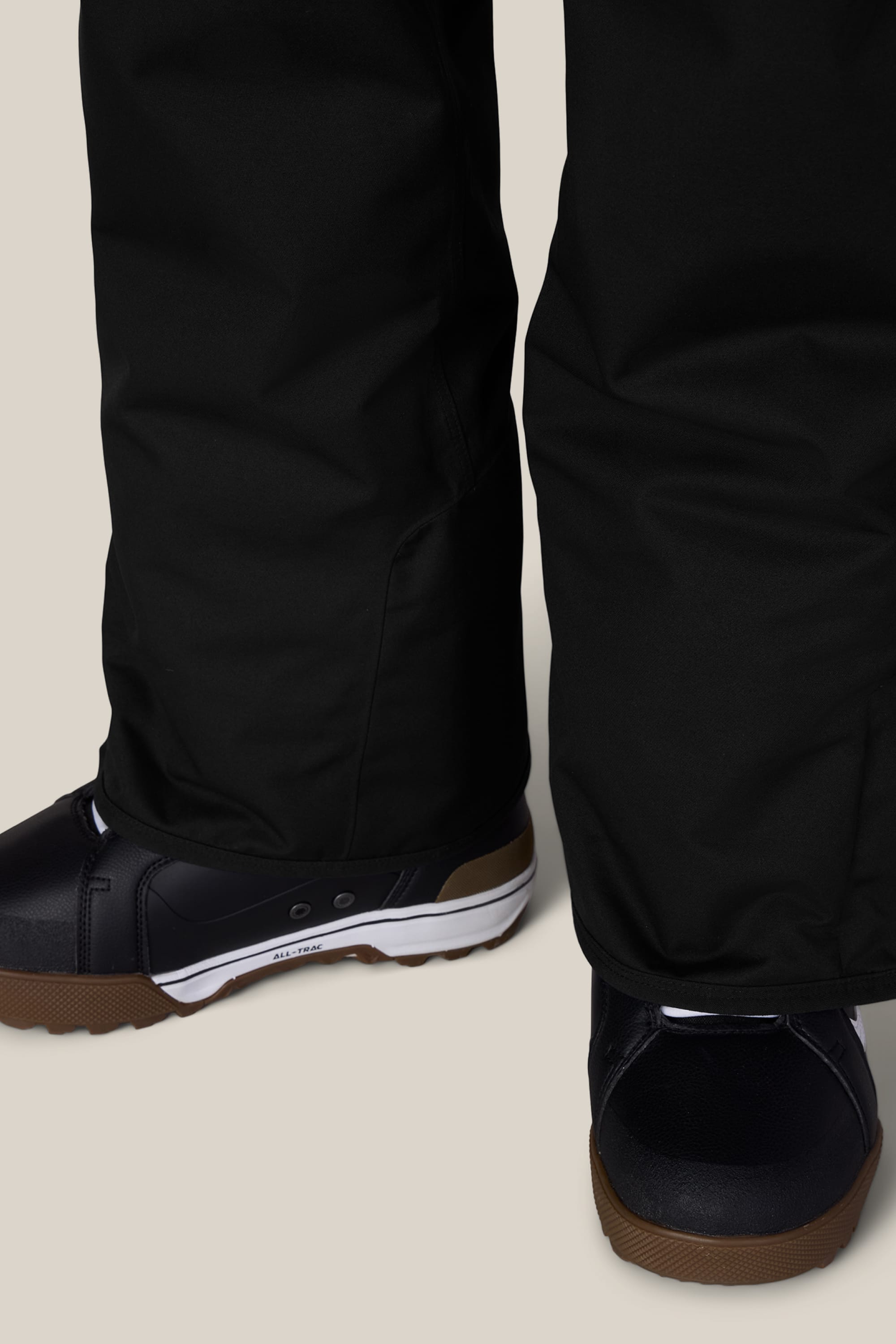 A close-up view of a person wearing the 686 Men’s Infinity™ Insulated Cargo Pant in black, paired with black sneakers featuring white midsoles and brown outsoles, standing on a light beige surface.