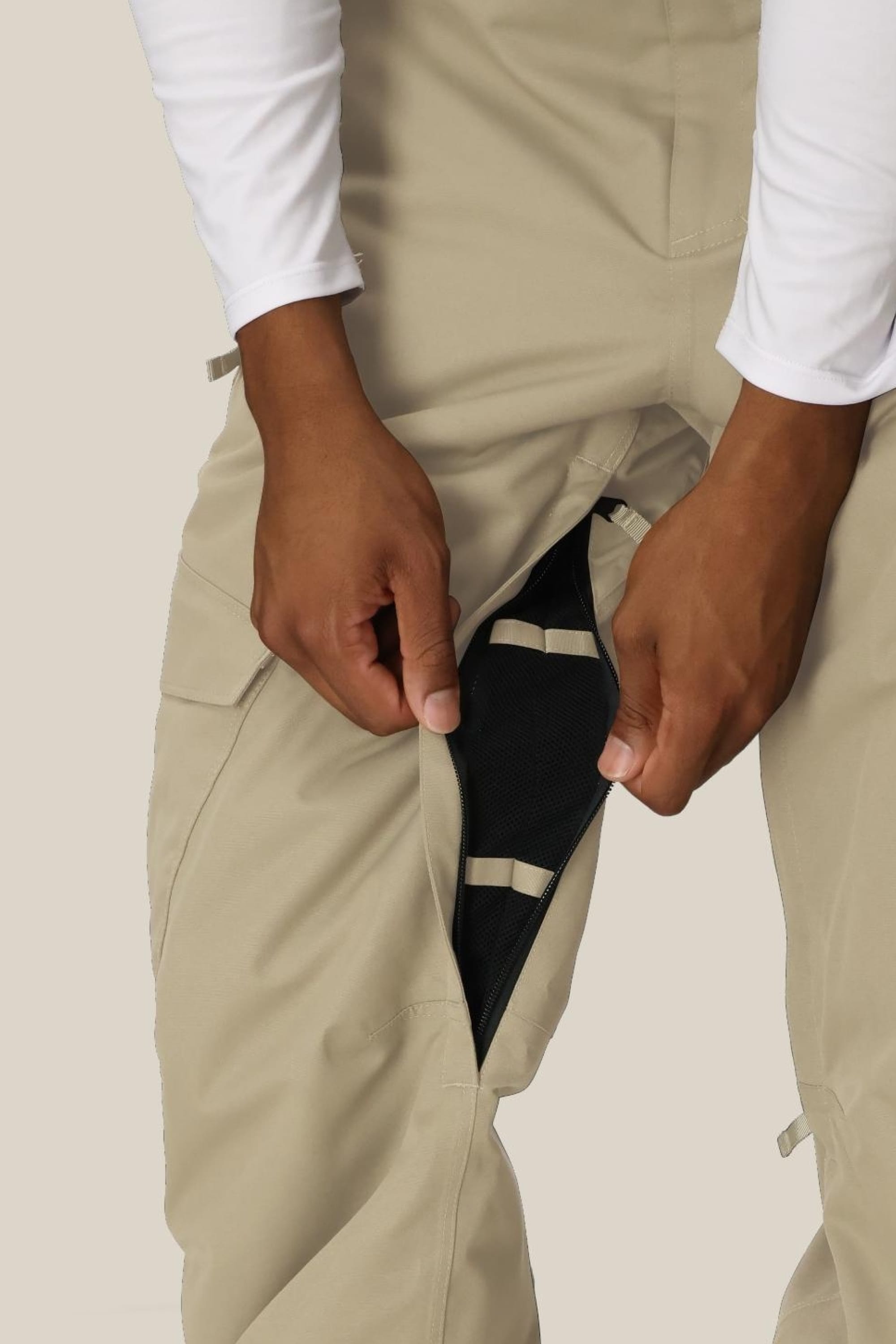 A person wearing the 686 Mens Infinity Insulated Cargo Pant unzips a leg vent to reveal black mesh fabric underneath. Their hands hold the zipper, and they are dressed in a white long-sleeve shirt.