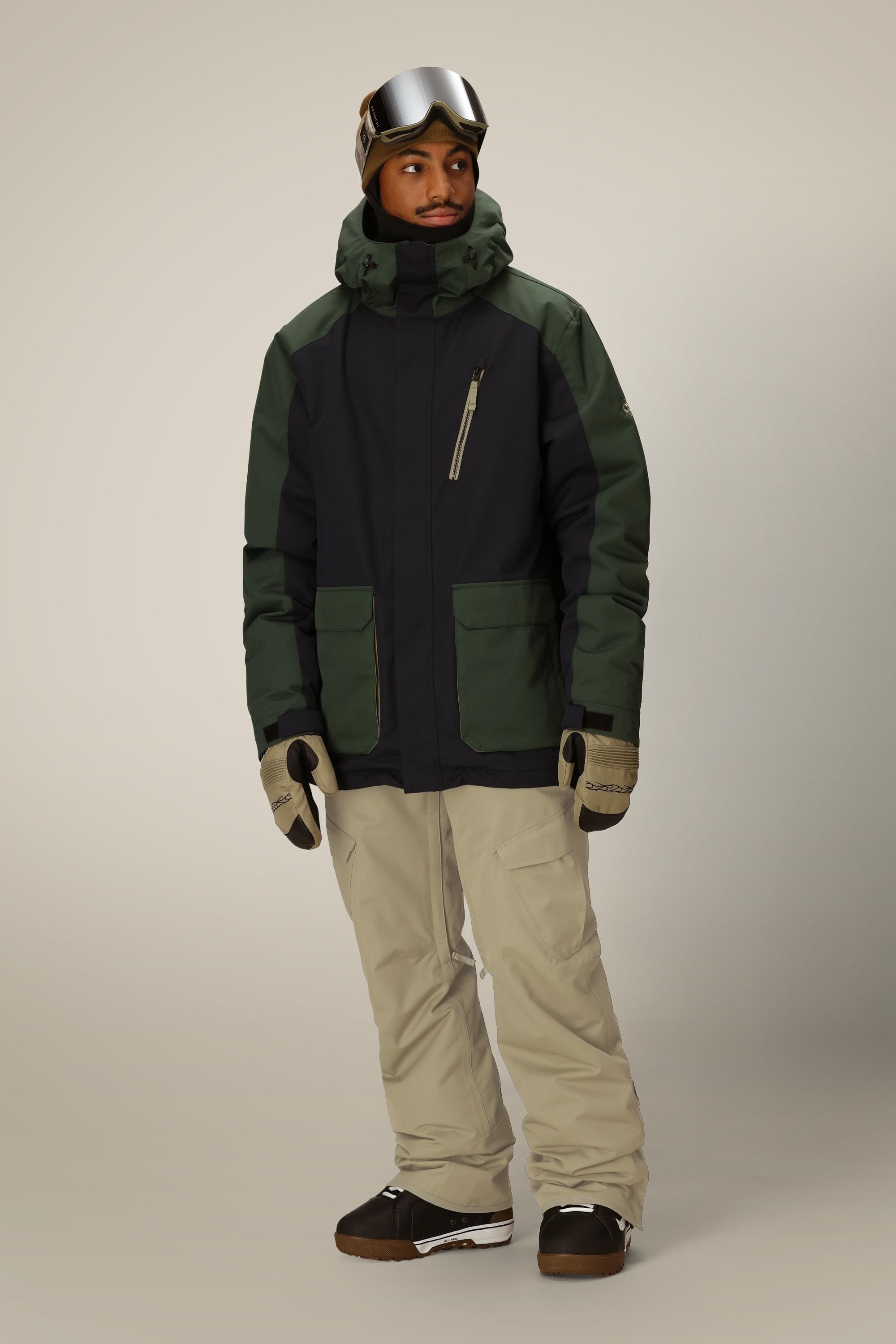 A person in a dark green and black winter jacket, brown boots, beige gloves, ski goggles on their head, and 686 Mens Infinity Insulated Cargo Pant by 686 stands against a plain light background.