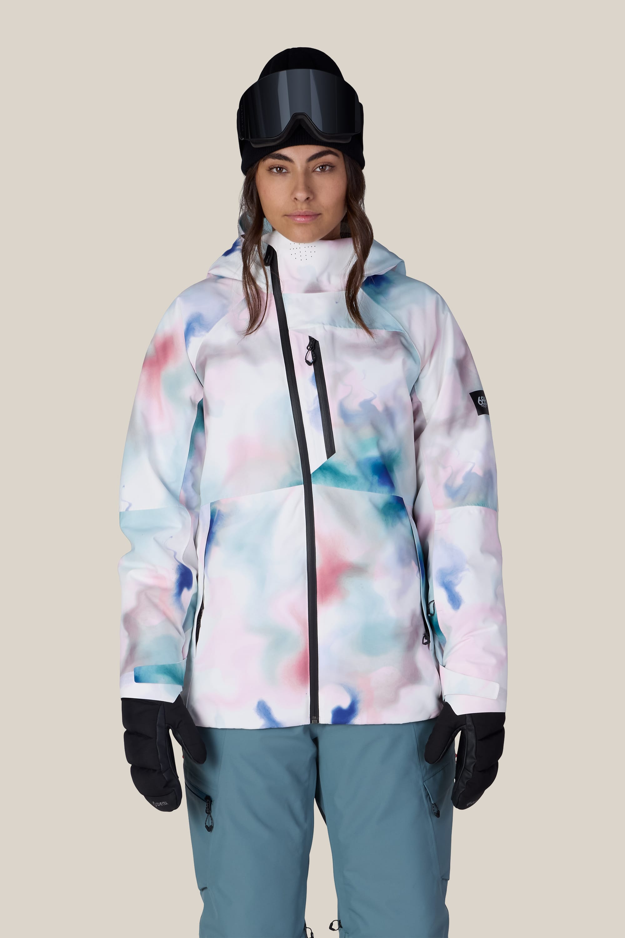 A woman wearing the 686 Women’s Hydra™ Thermagraph® Jacket by 686—featuring a colorful pastel pattern—along with blue snow pants, black gloves, and a black helmet with goggles, stands against a plain light background.