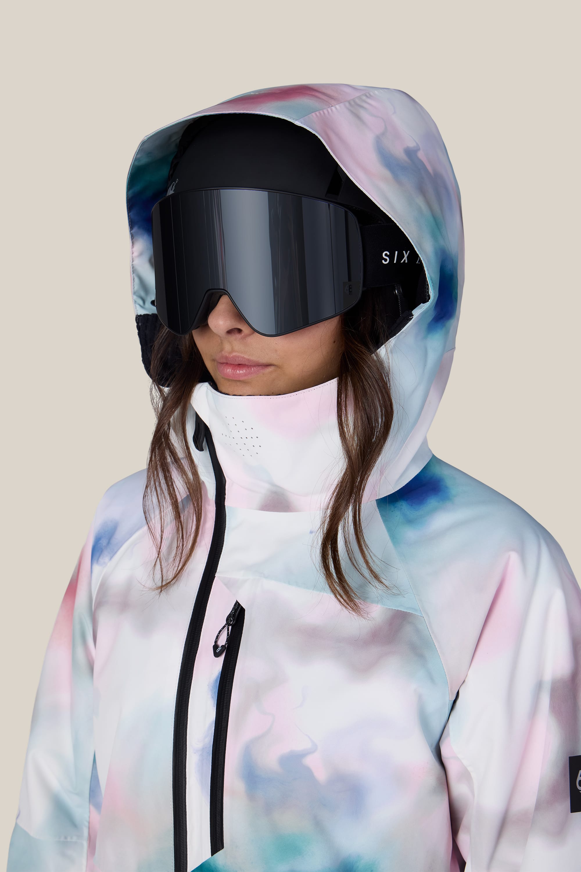 A person wearing the 686 Women’s Hydra™ Thermagraph® Jacket by 686, paired with large black ski goggles and a face covering, stands against a neutral beige background.